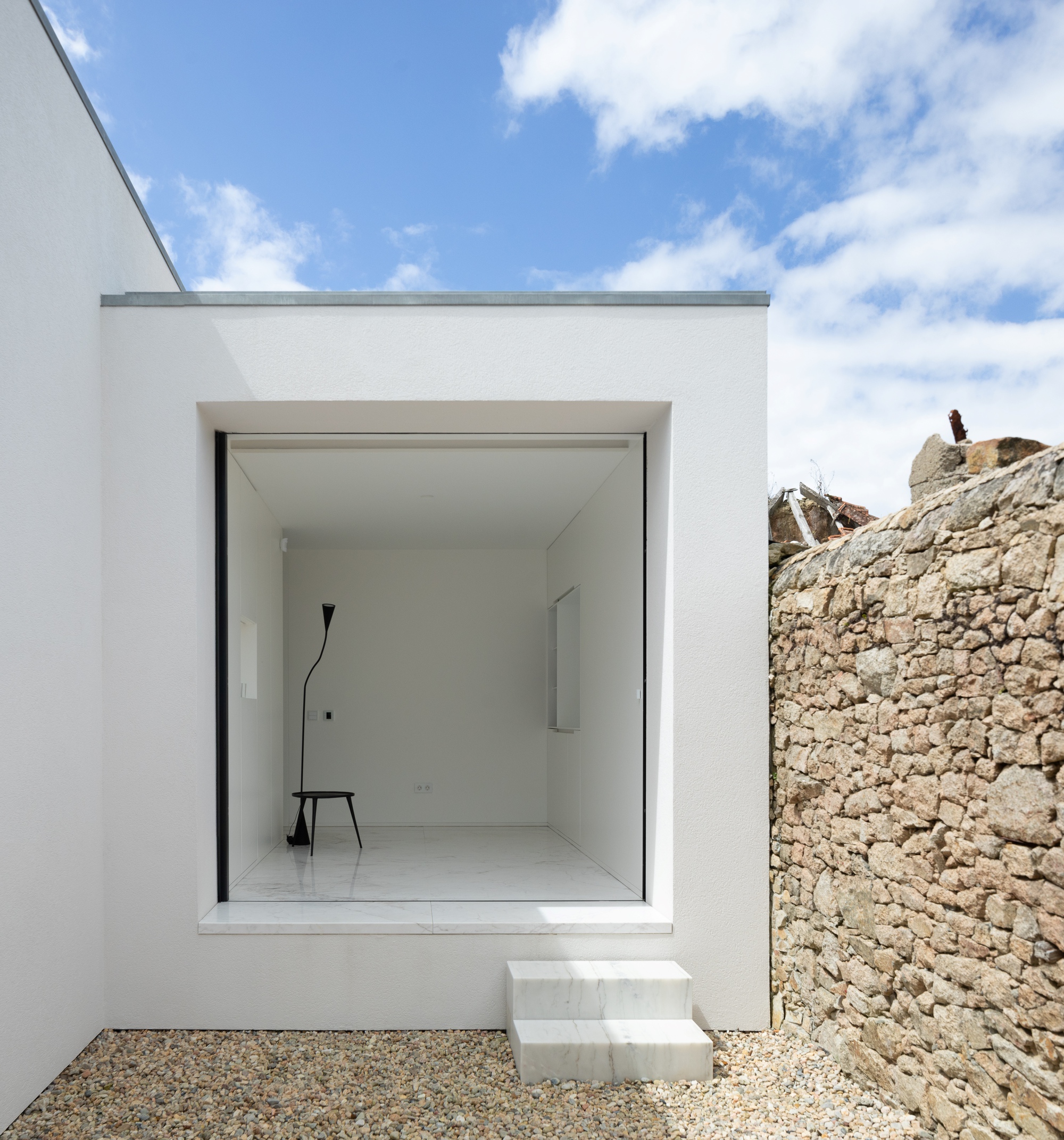 Deep square opening in white wall holding a black stool and floor lamp, with marble steps beside a rough stone wall