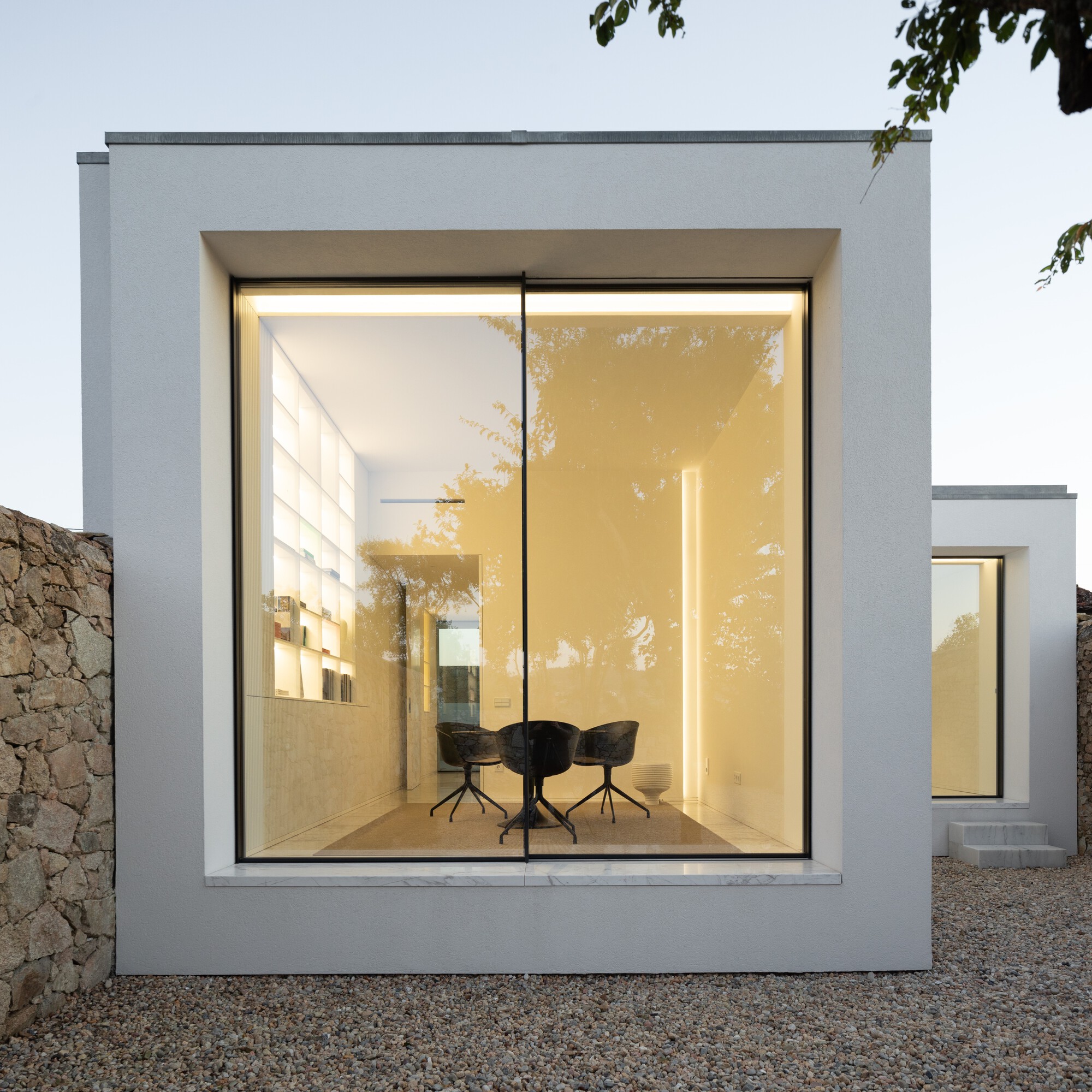 White stucco box with a deep frame around a floor-to-ceiling glazed dining room, glowing at dusk beside a dry stone wall