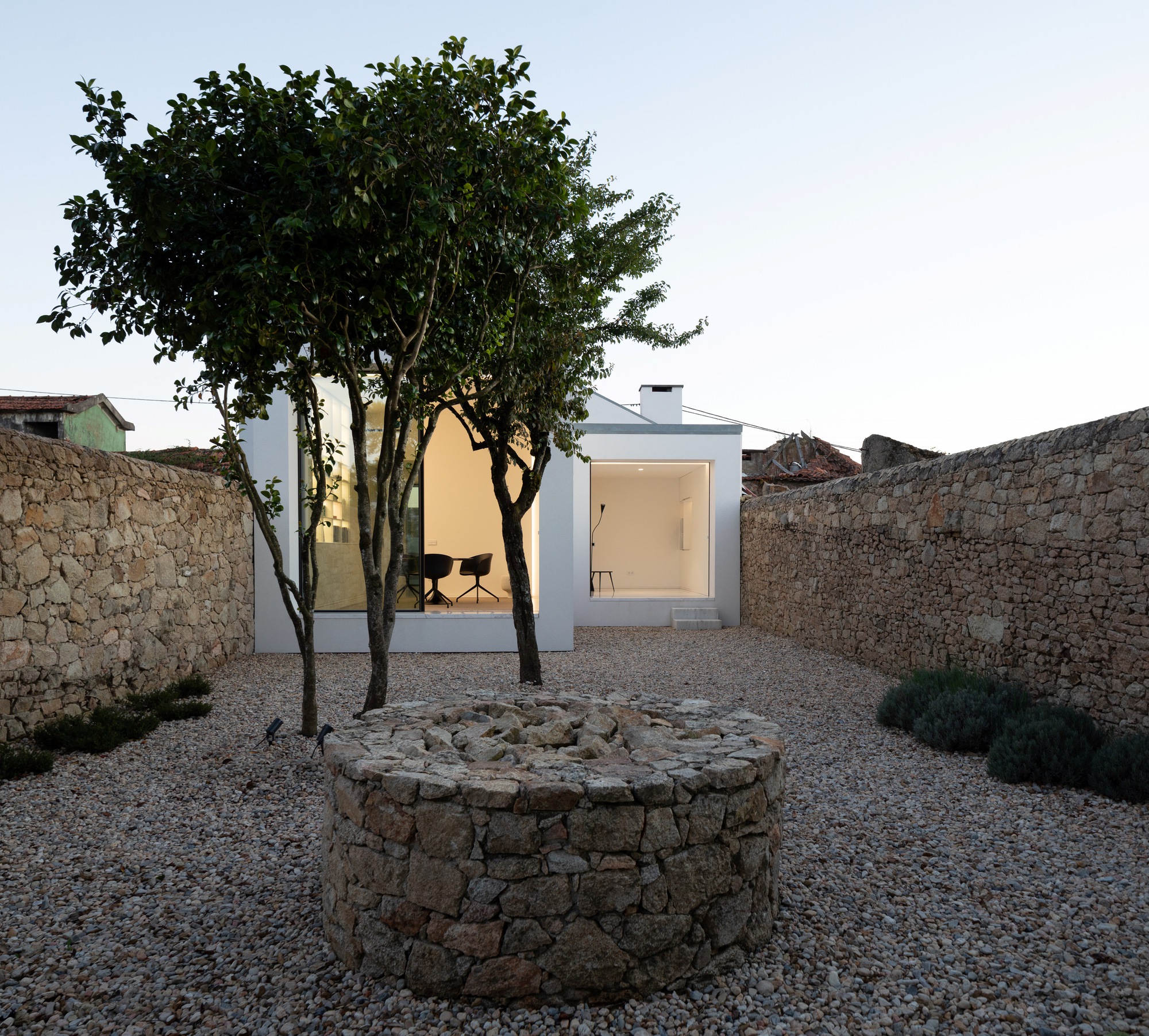 Gravel courtyard with a round stone well and two slender trees in front of the glazed white volumes at dusk