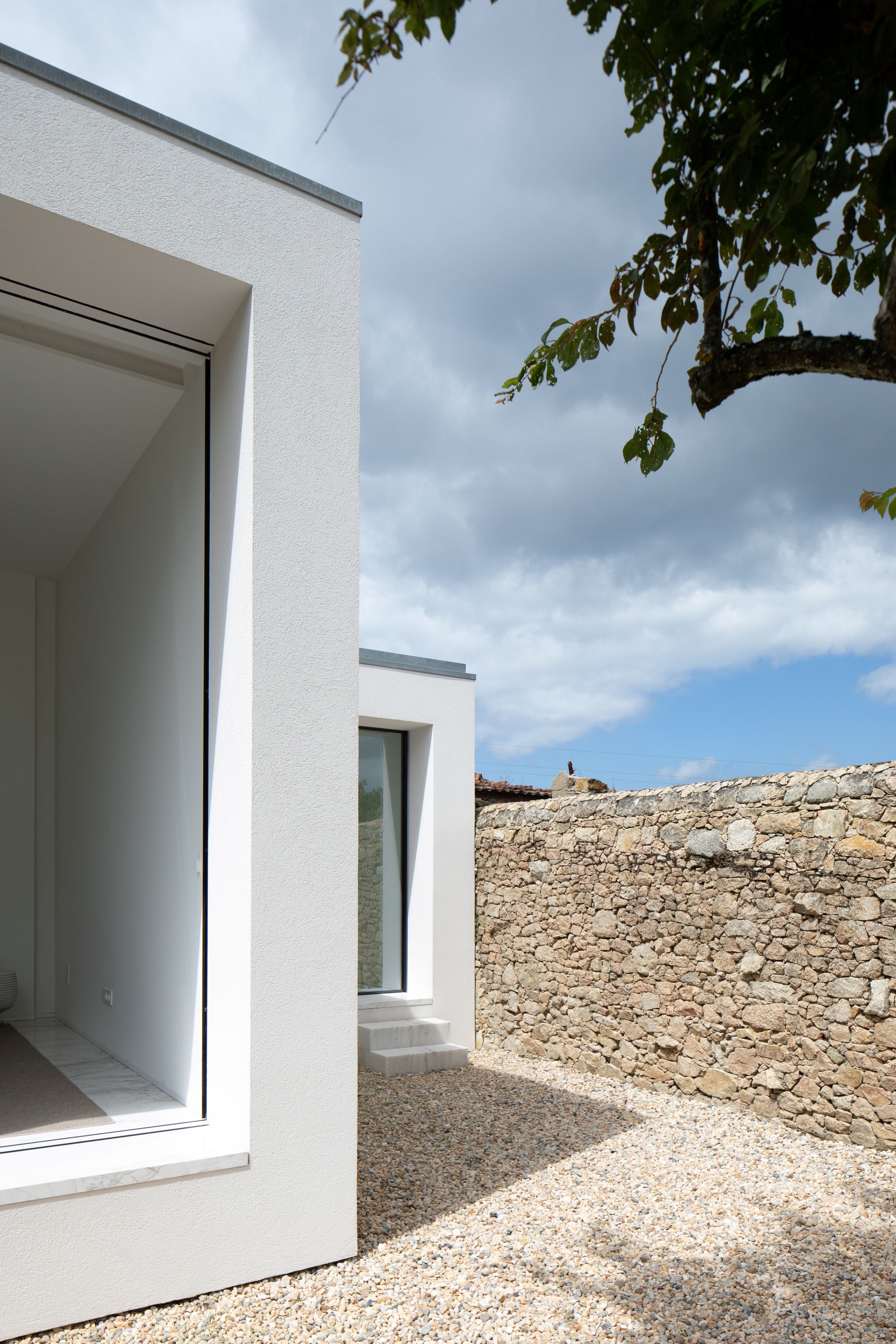 Corner of the white stucco volumes with recessed glazing and a marble sill, beside a stone wall under cloudy sky