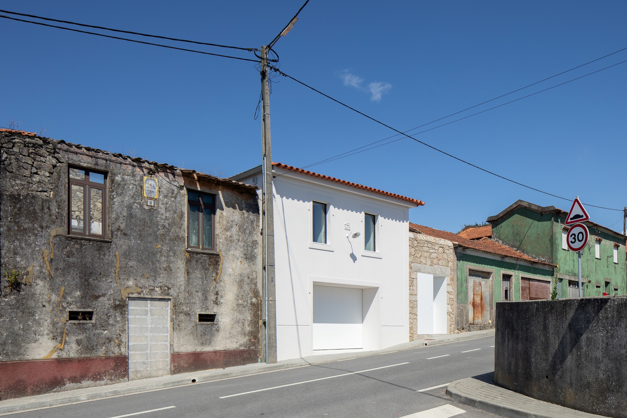 Street view of the white house with terracotta roof between weathered stone and green-painted neighbours, under a clear blue sky