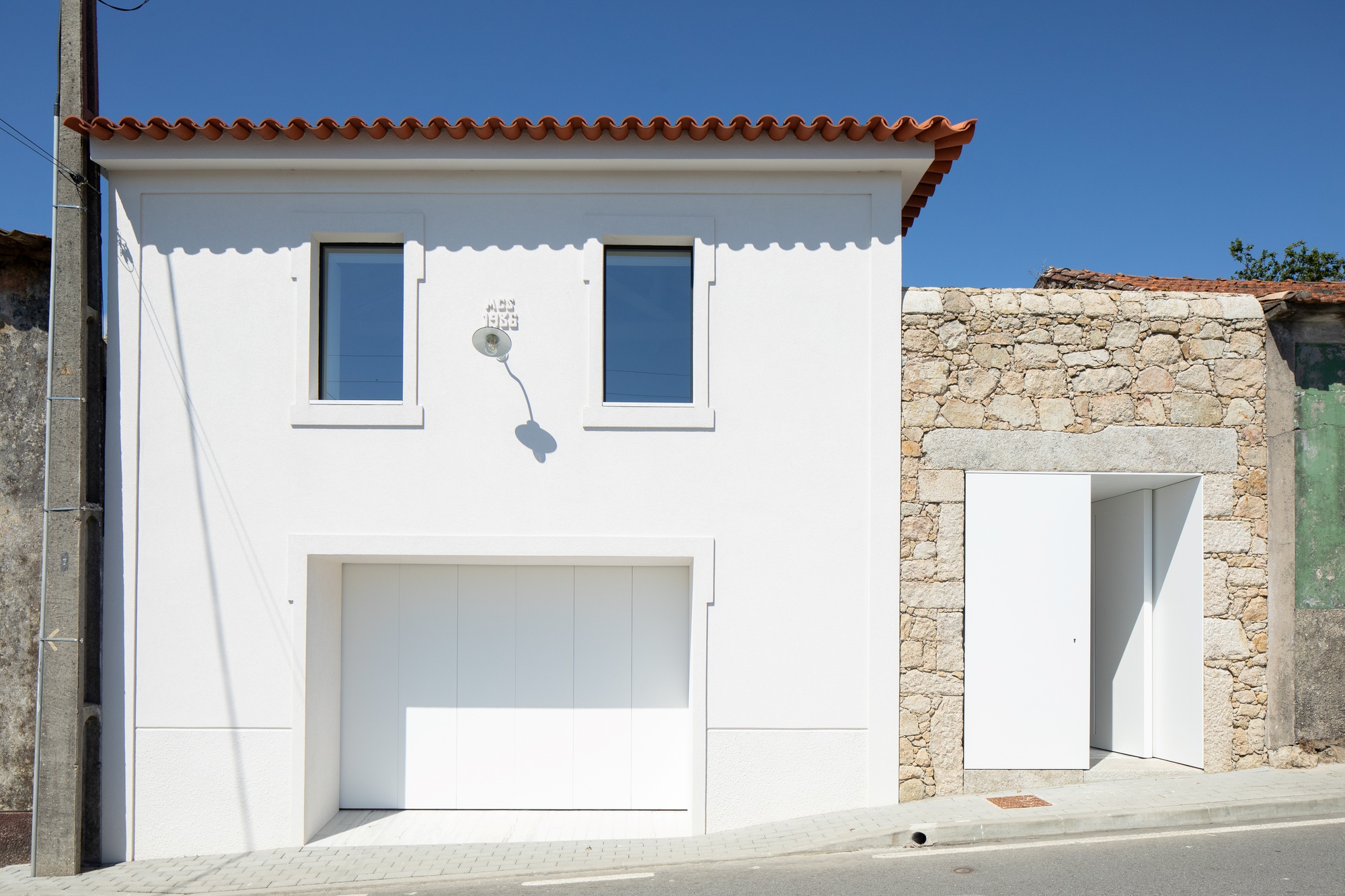 Street facade with two framed windows, white garage panel door and a stone wall with open pedestrian door in midday sun