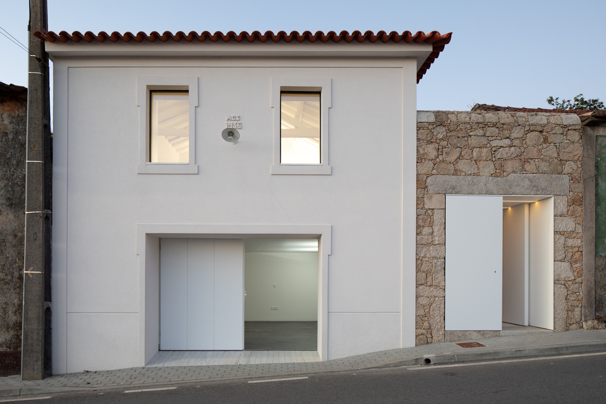 Street facade at dusk with the garage door slid open and warm light inside the upper windows and stone-framed entry