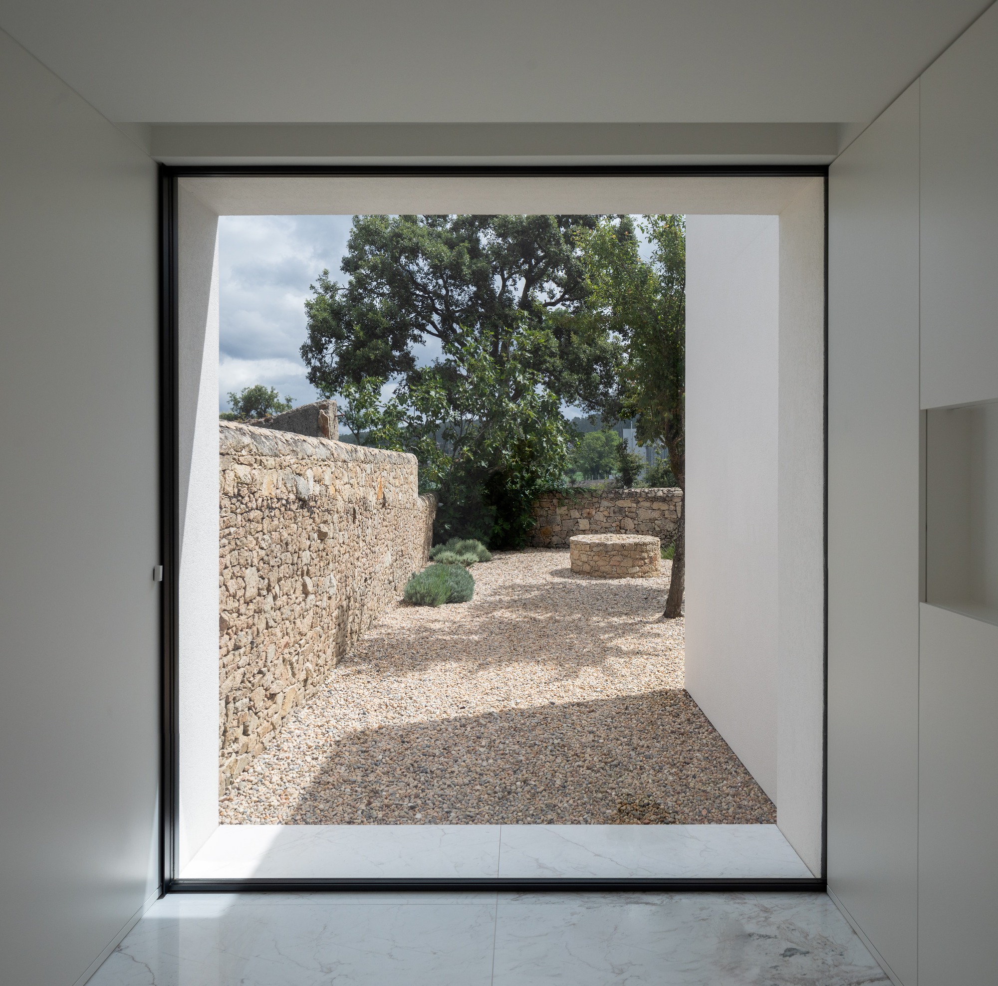 Framed view from a white interior through a floor-to-ceiling window onto a gravel courtyard, dry stone wall and oak tree under cloudy skies