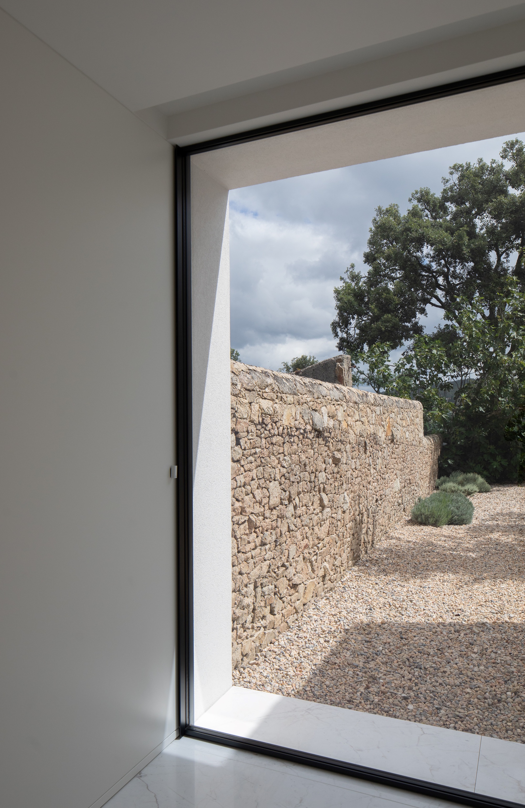 Side view through a tall black-framed window toward a dry stone wall and lavender bushes on gravel, with clouds above