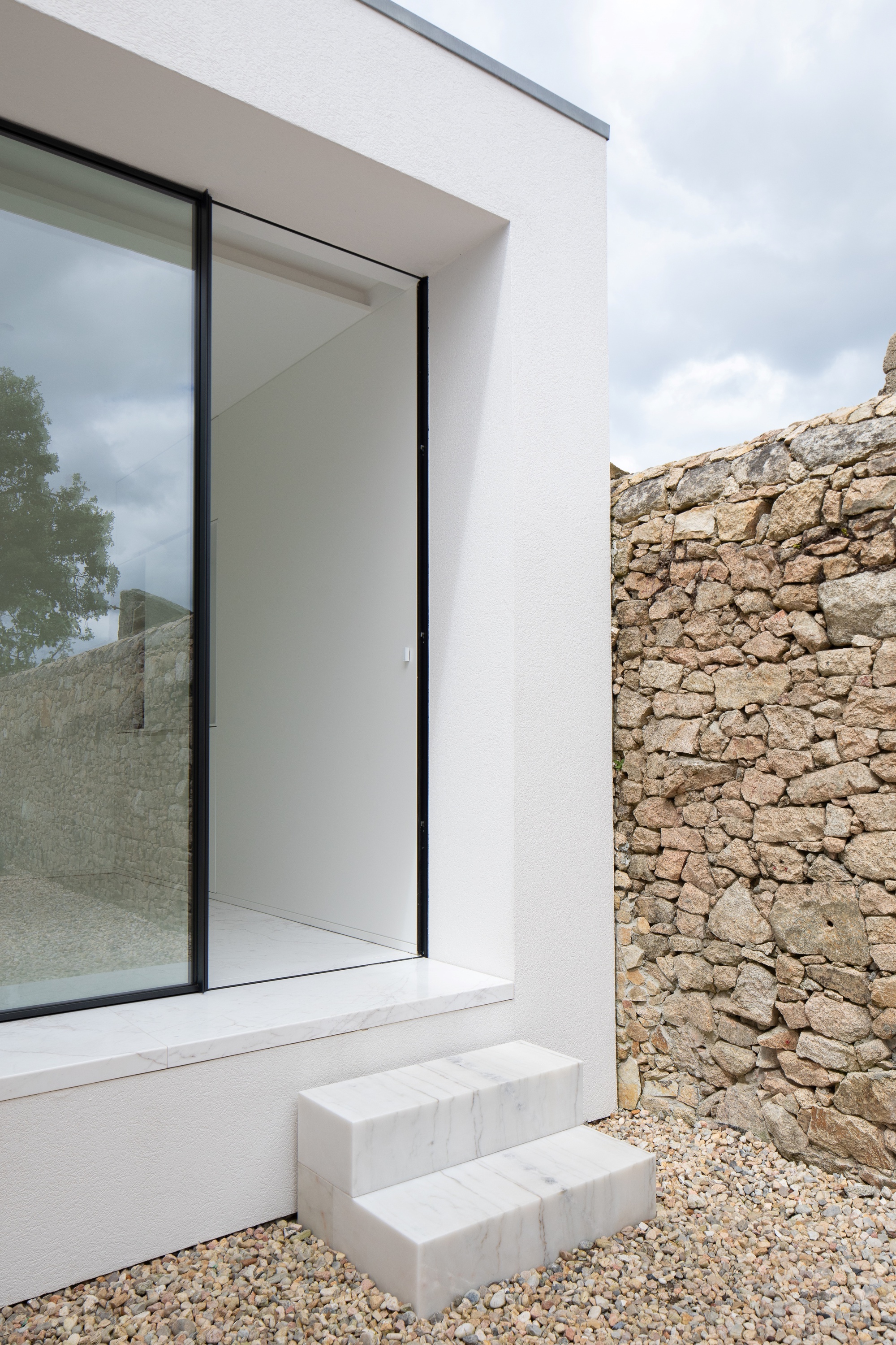White rendered volume with a recessed glazed opening, marble sill and two marble steps beside a rough stone wall on gravel