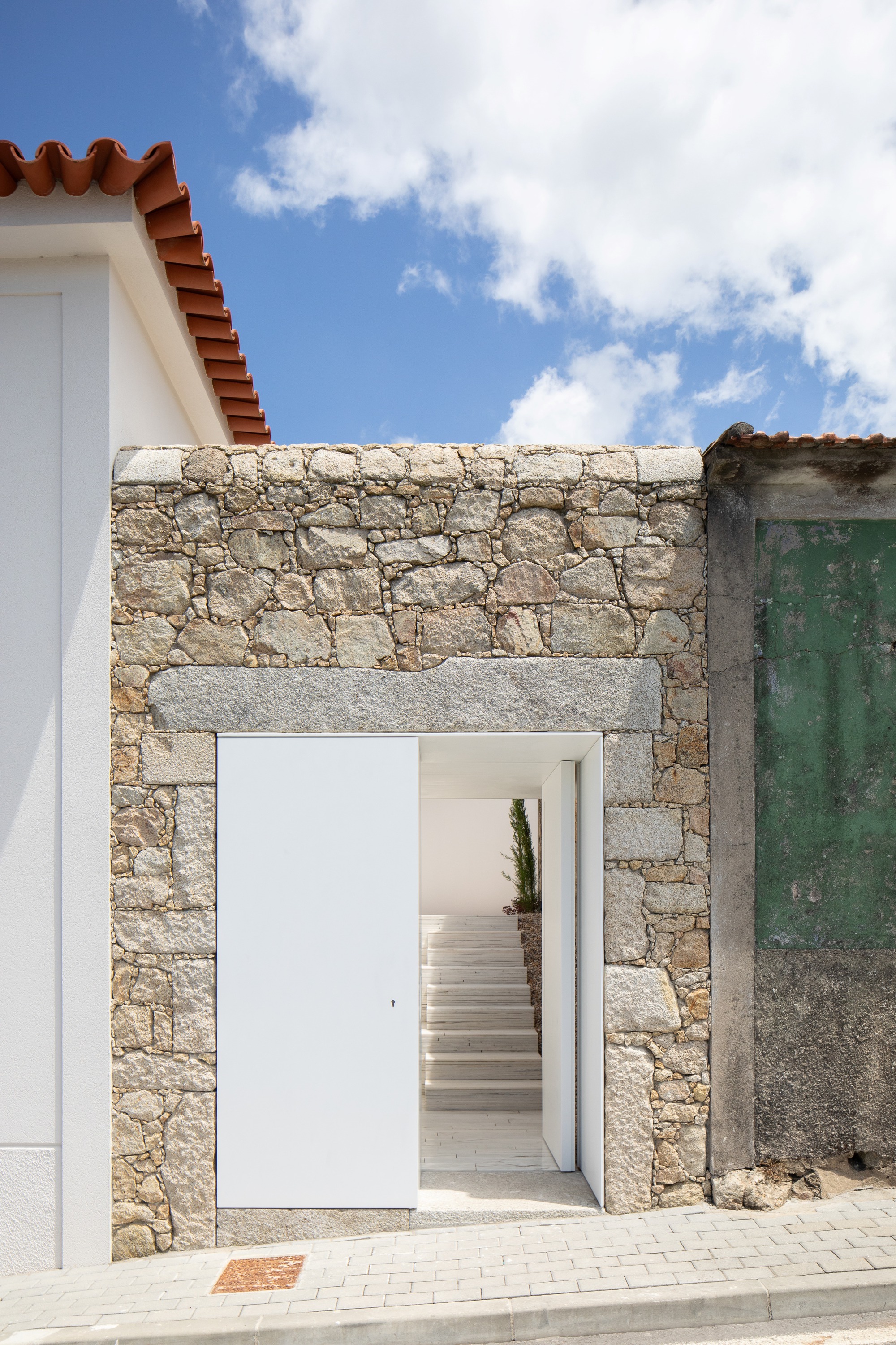 Street entrance in a rubble stone wall with a granite lintel and pivoting white door, opening to steps, under a blue sky