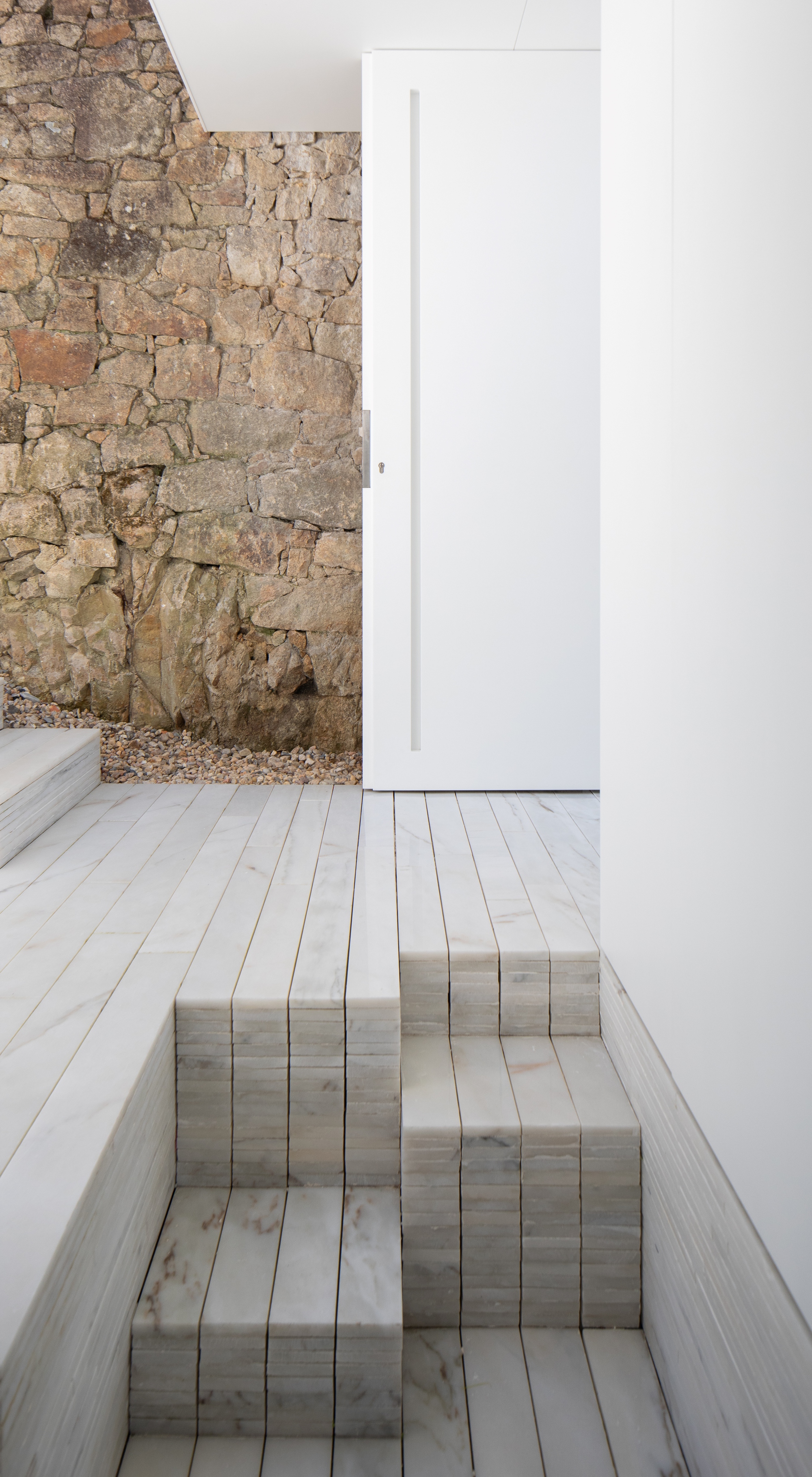 Pale marble strip steps rising to a tall white pivot door beside a rough stone retaining wall and gravel edge