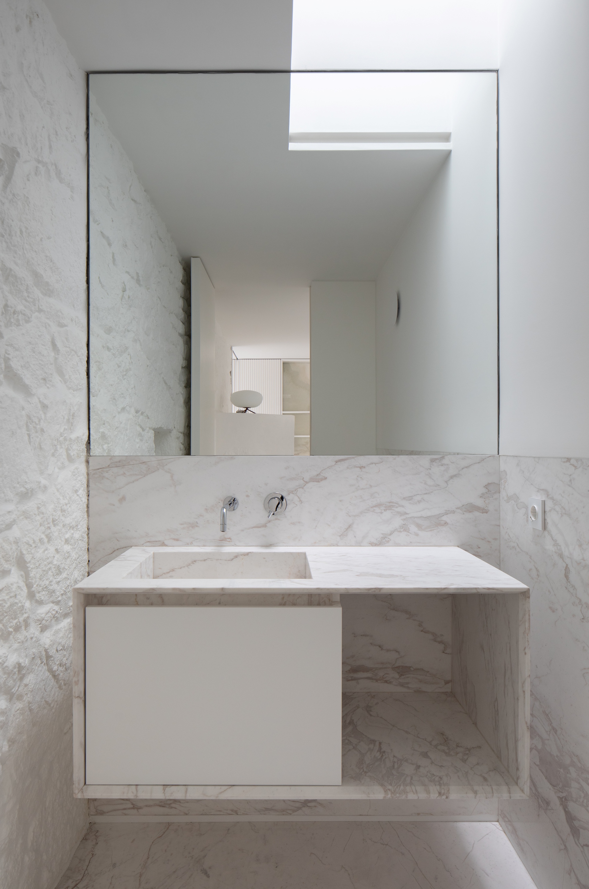 Floating marble vanity with wall-mounted taps beneath a large mirror, reflecting a skylight and a white rough-plastered wall