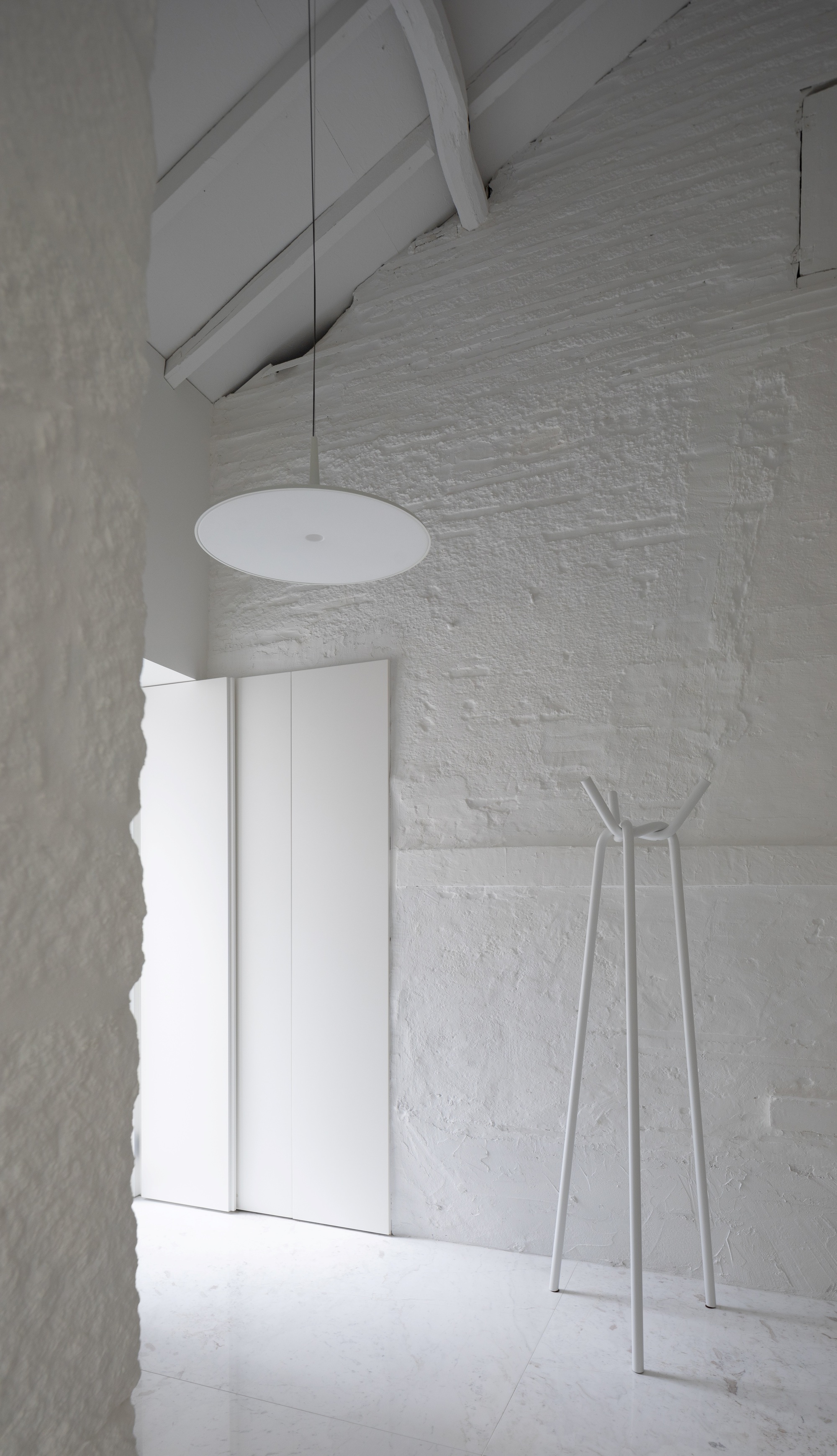 Whitewashed brick wall under exposed roof timbers, with a disc pendant lamp and a slim white coat stand