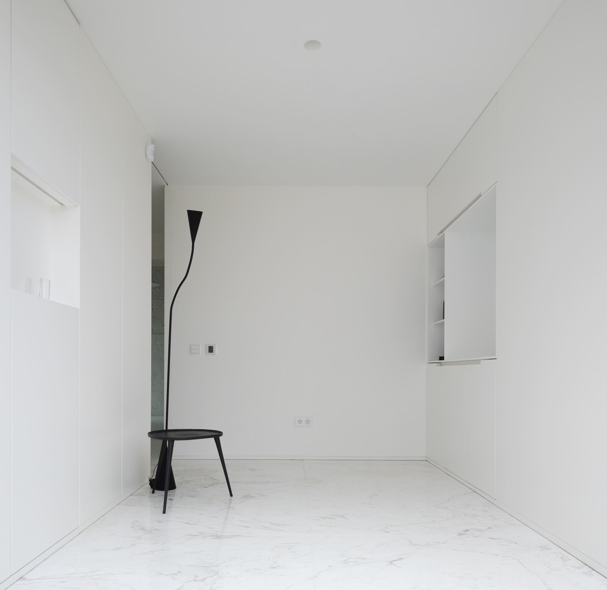 Empty white room with flush cabinetry, a black tripod side table with a curved floor lamp, and a marble floor