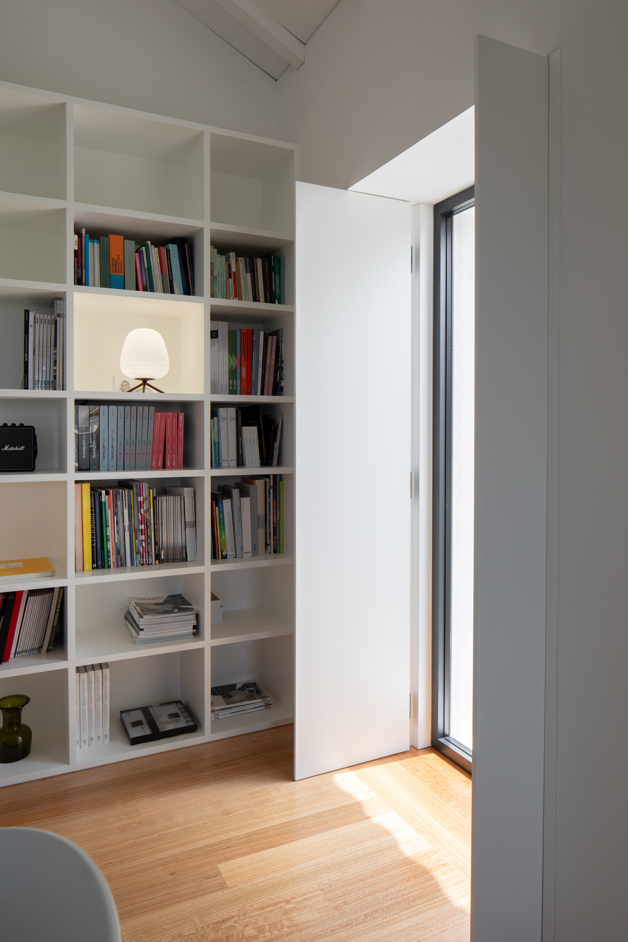 White built-in bookshelf beside a flush door and tall window, with sunlight falling across the oak floorboards