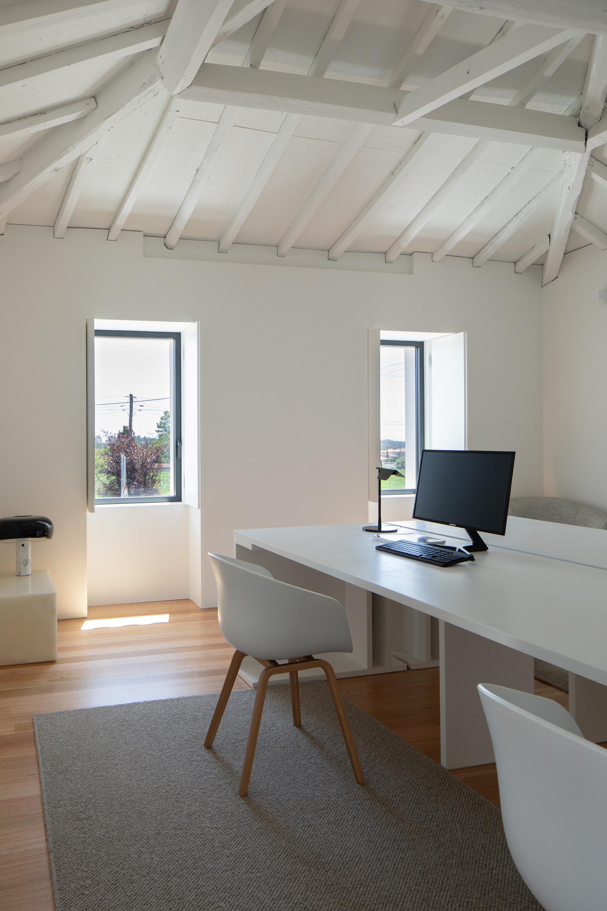 Attic workspace with white exposed roof rafters, a long desk and two small windows looking onto trees and power lines
