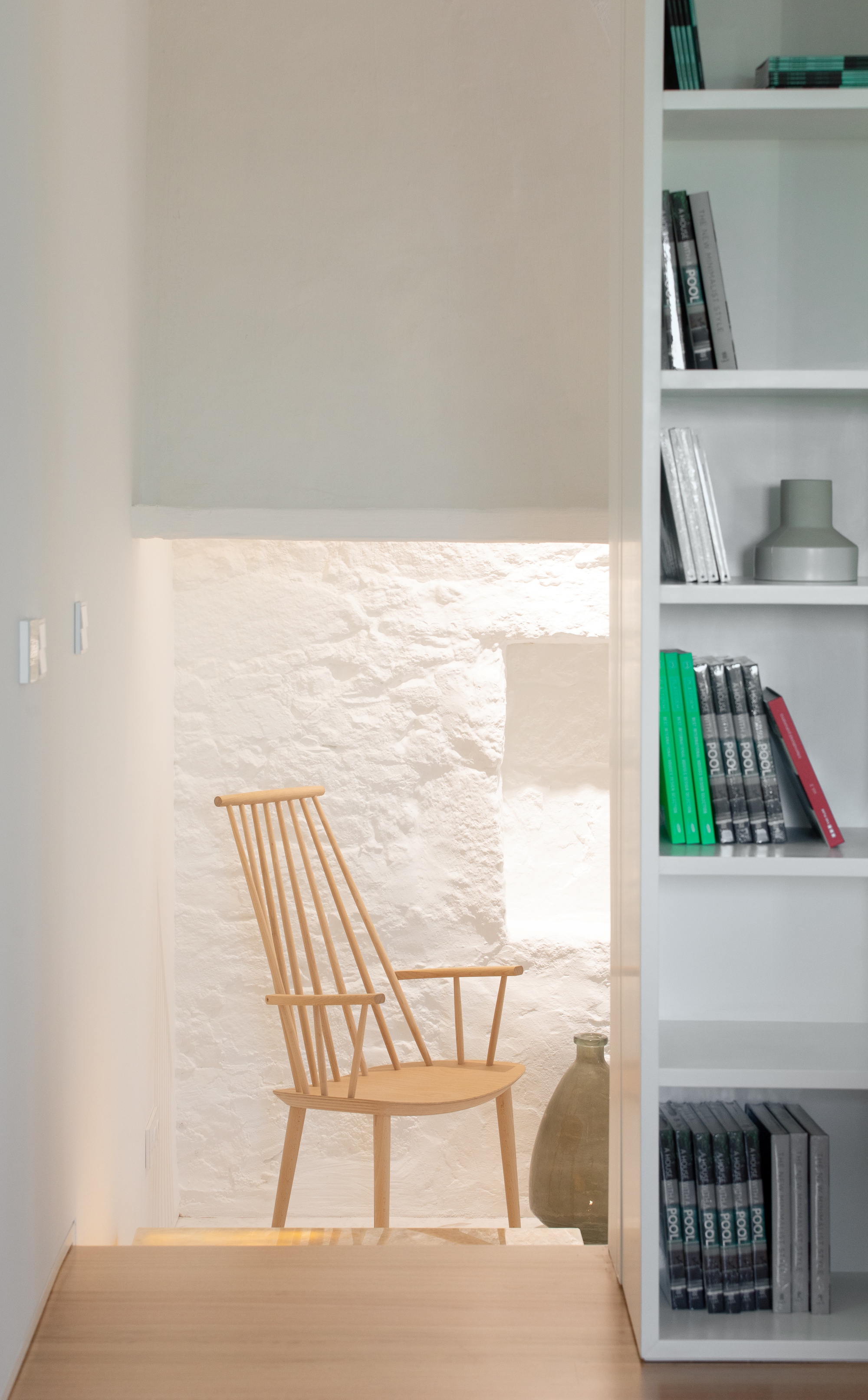 Wooden spindle-back chair in a lit whitewashed stone recess, seen past a white bookshelf holding green and grey spines