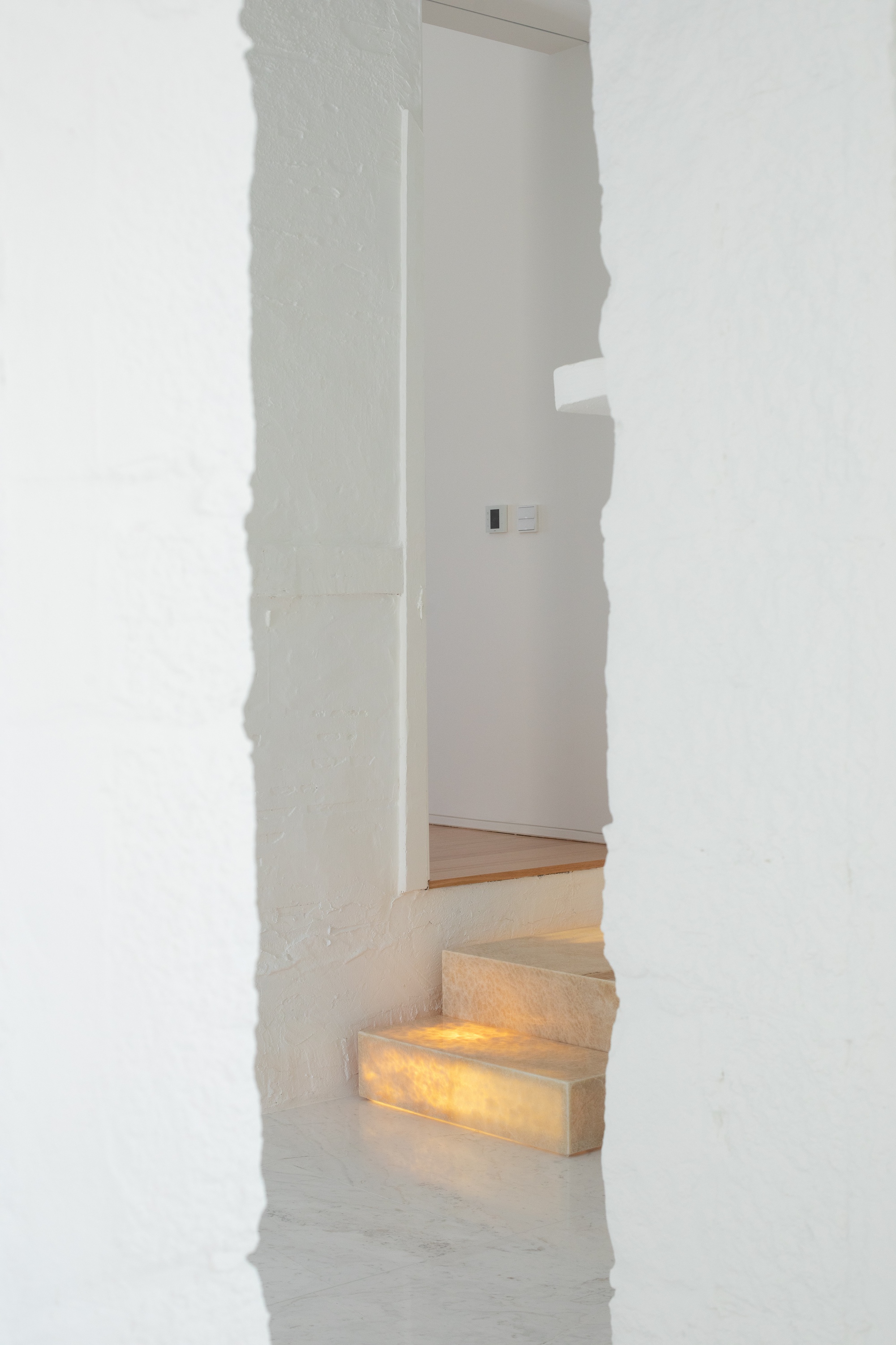 Narrow gap in a rough whitewashed wall framing backlit onyx steps and a corridor with a light switch beyond