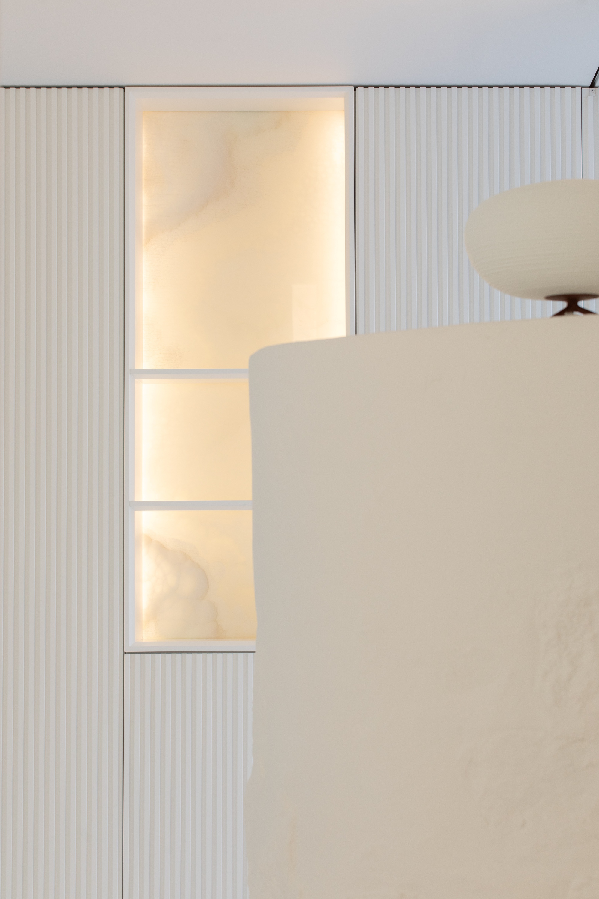 Backlit onyx panel set into a fluted white wall, with a rounded plaster edge and a globe lamp in the foreground