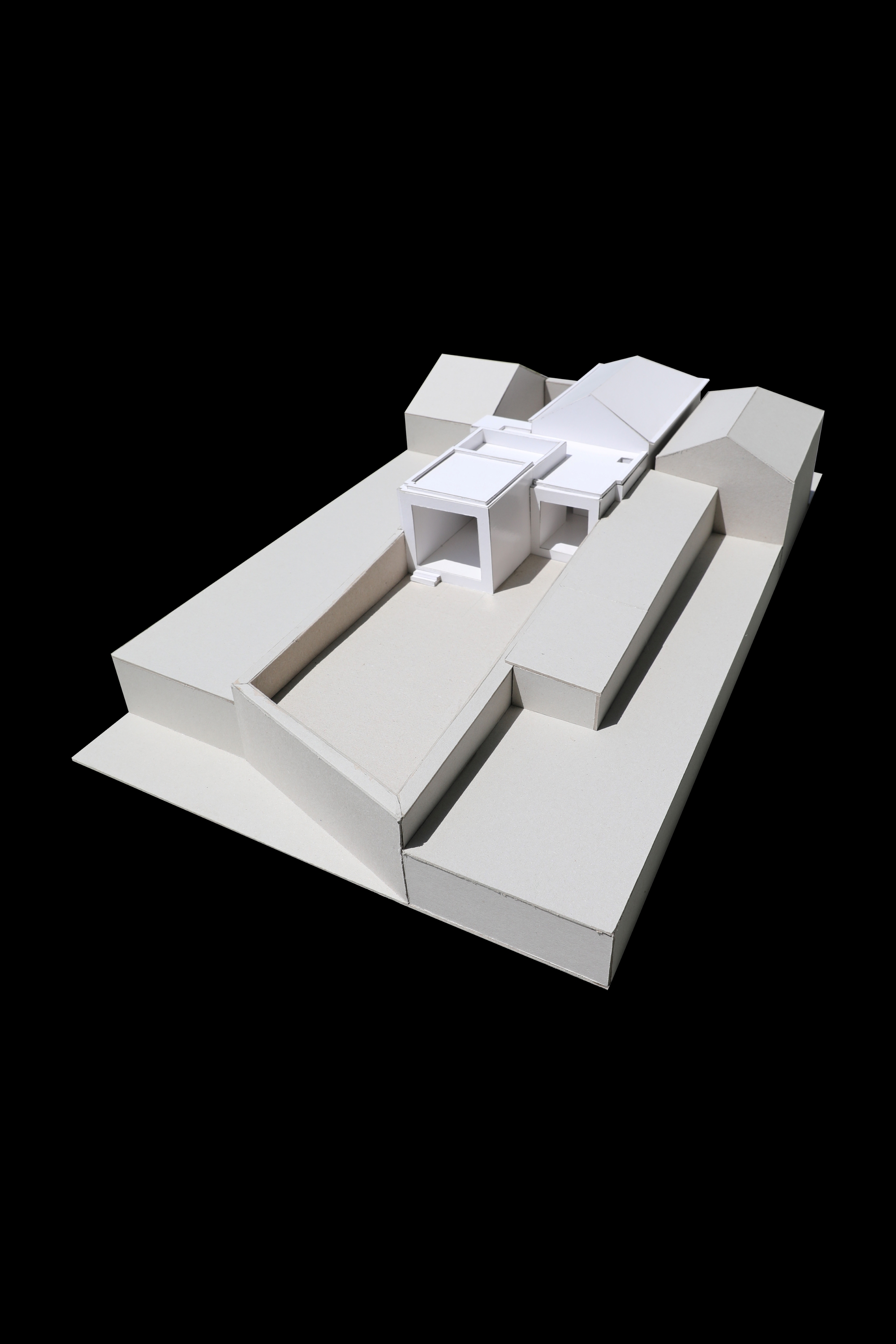 White card massing model seen from a high corner, with a small boxy volume set between a gabled house and a long terrace wall in hard sunlight