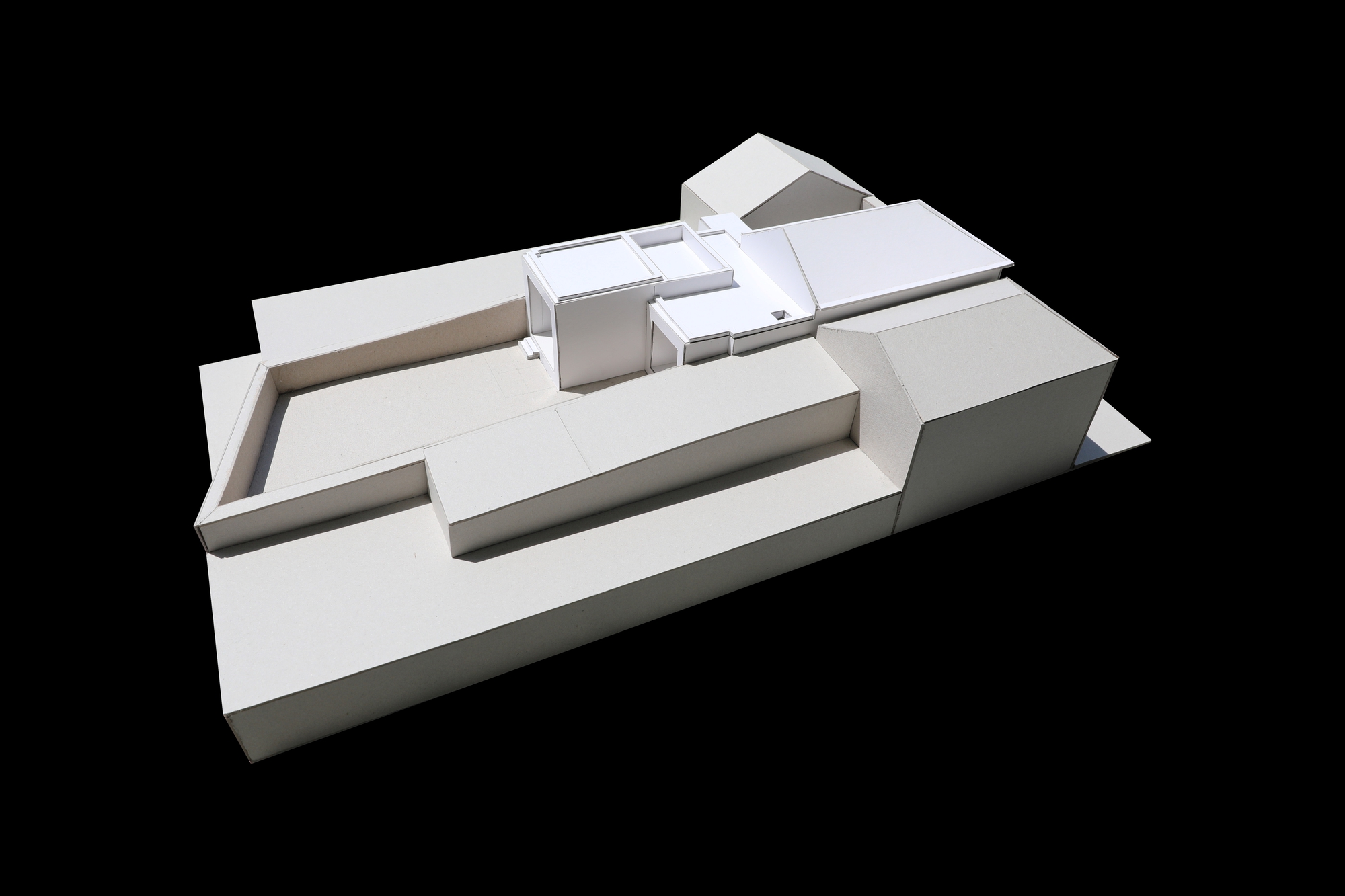 White card site model from the long side, showing a walled terrace, the central flat-roofed volumes and a gabled block, with sharp shadows on black