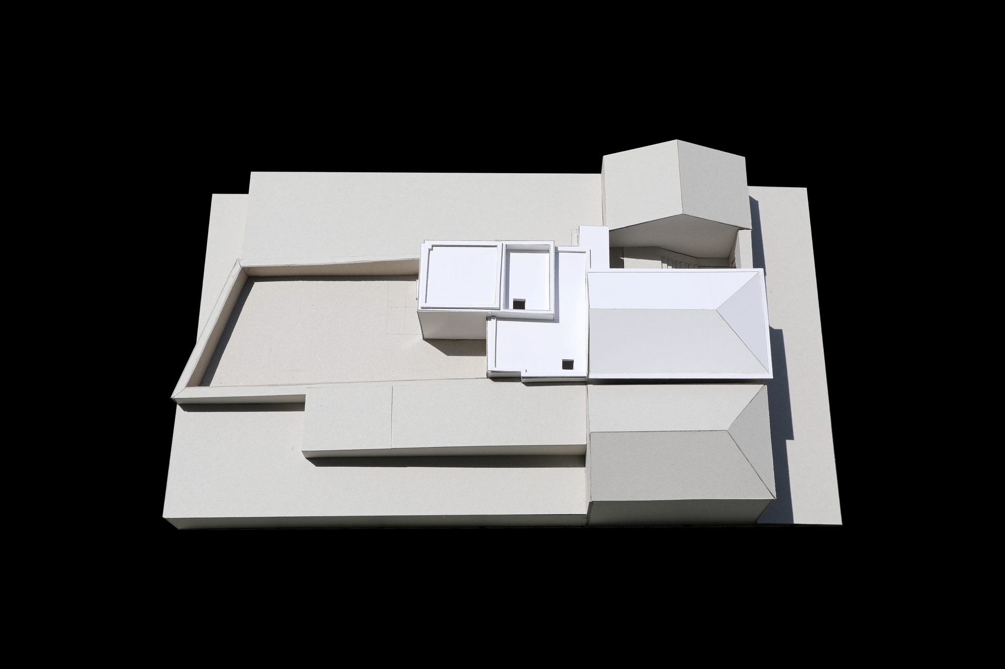 Top-down view of the white card model showing flat roofs with small openings beside a hipped roof and a terrace wall, in direct sun