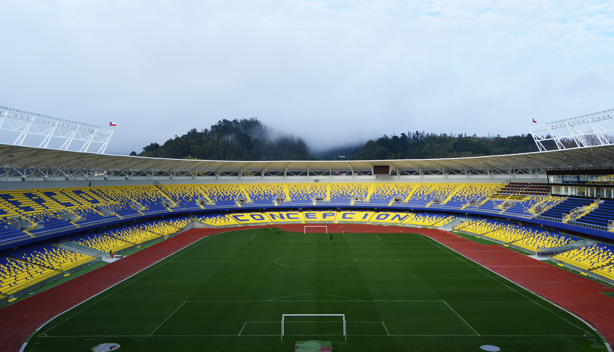 Pitch seen from the stands with red running track, yellow and blue seating and a mist-covered wooded hill beyond the roof edge