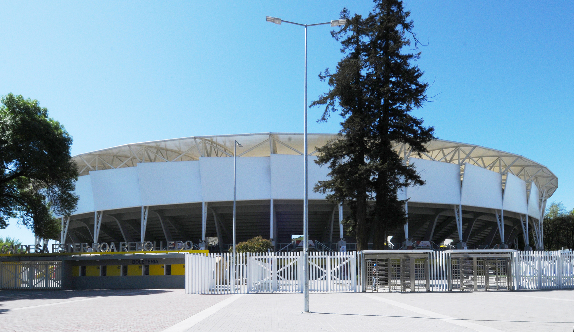 Exterior of the oval stadium with white panel facade and steel roof trusses behind a tall pine and lamp post under clear sky