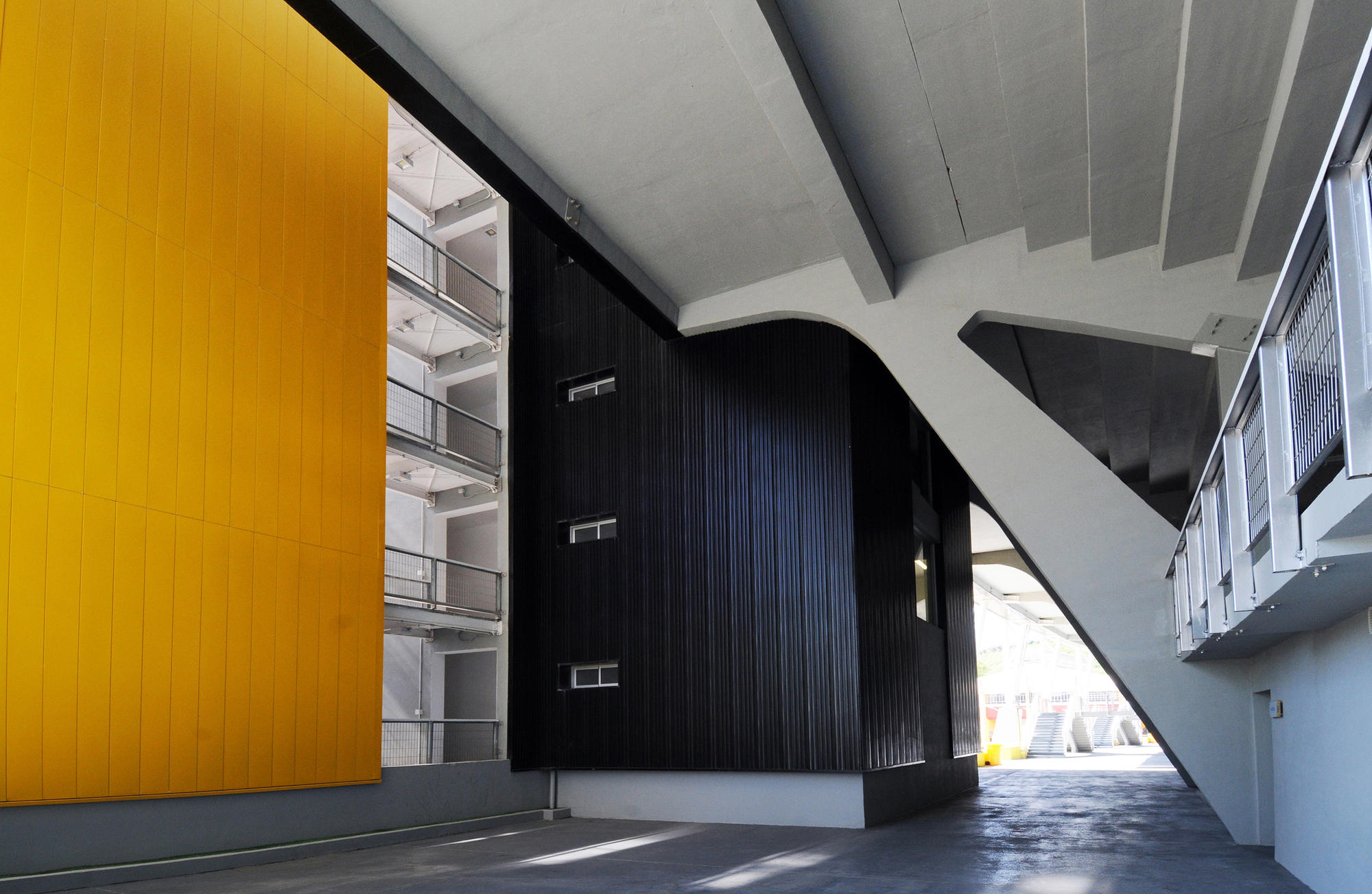 Concourse beneath the stands where a yellow panel wall, black clad volume and sloping concrete support meet in afternoon light