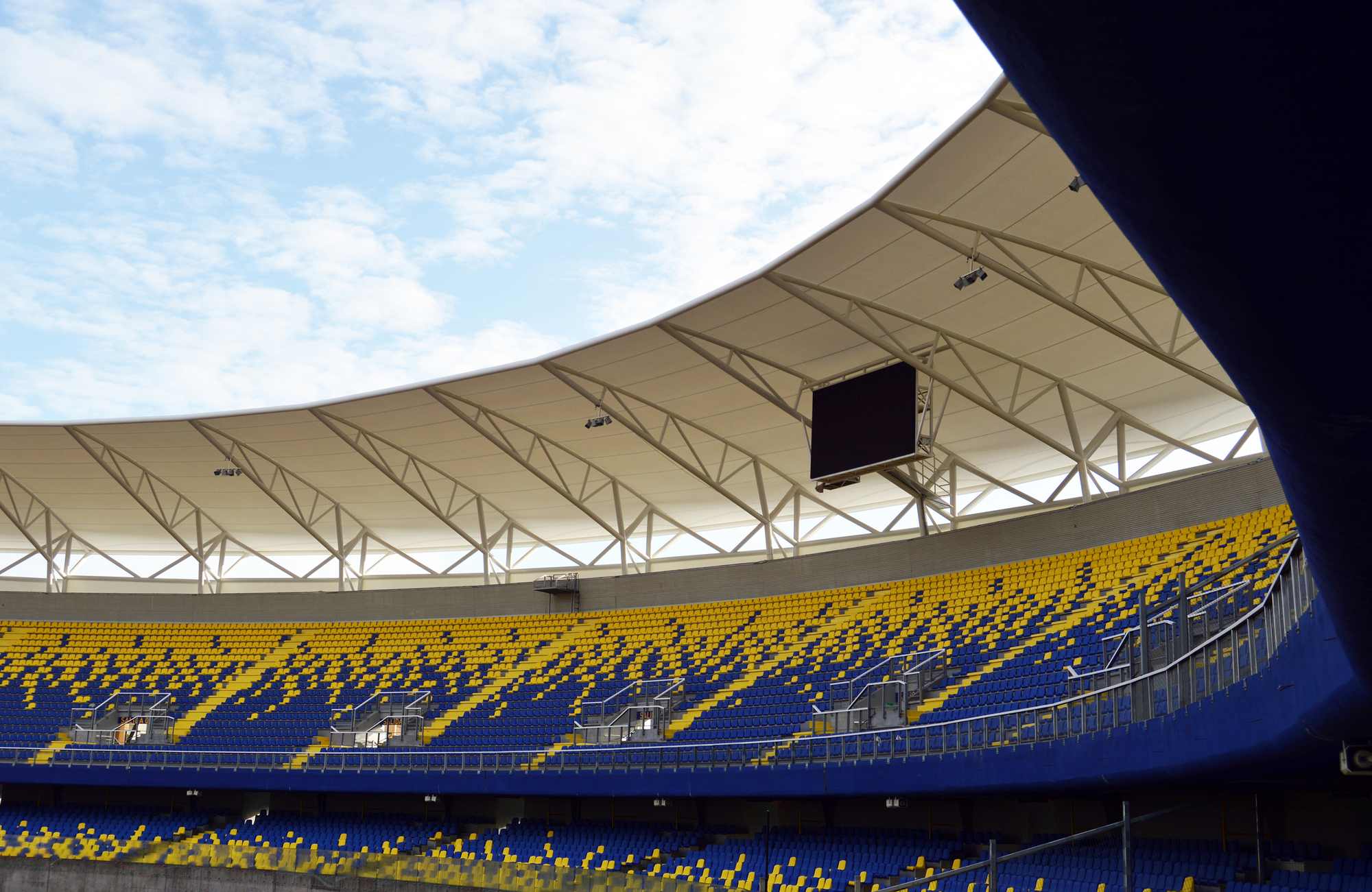 Tension roof with white steel trusses curving over yellow and blue seating, with a video screen hanging beneath a cloudy sky