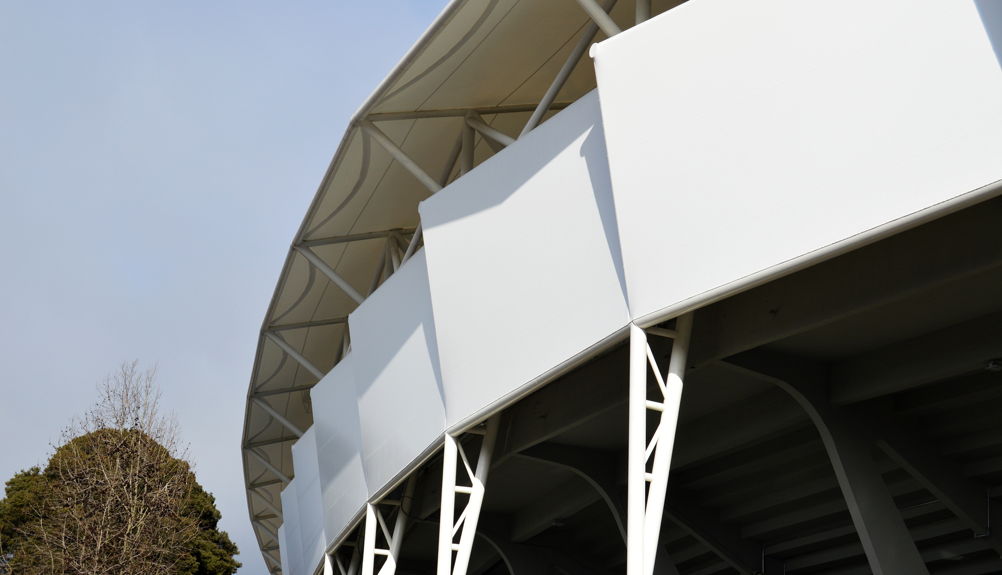Close-up of white facade panels held by lattice steel brackets beneath the roof edge, with a bare tree in the corner