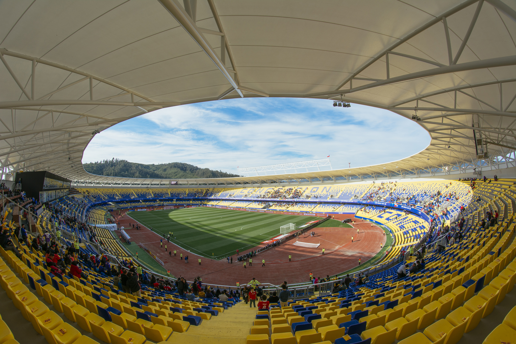 Fisheye view from the upper seating of the oval roof opening, pitch and red track, with spectators gathered on a sunny day
