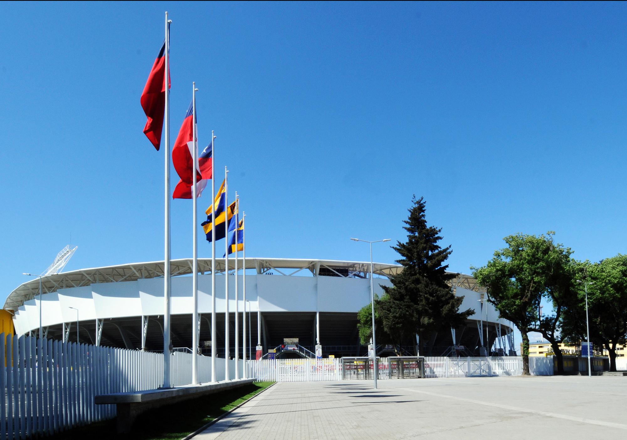 White stadium facade with a curved canopy roof beyond a row of flagpoles and a paved plaza under clear blue sky