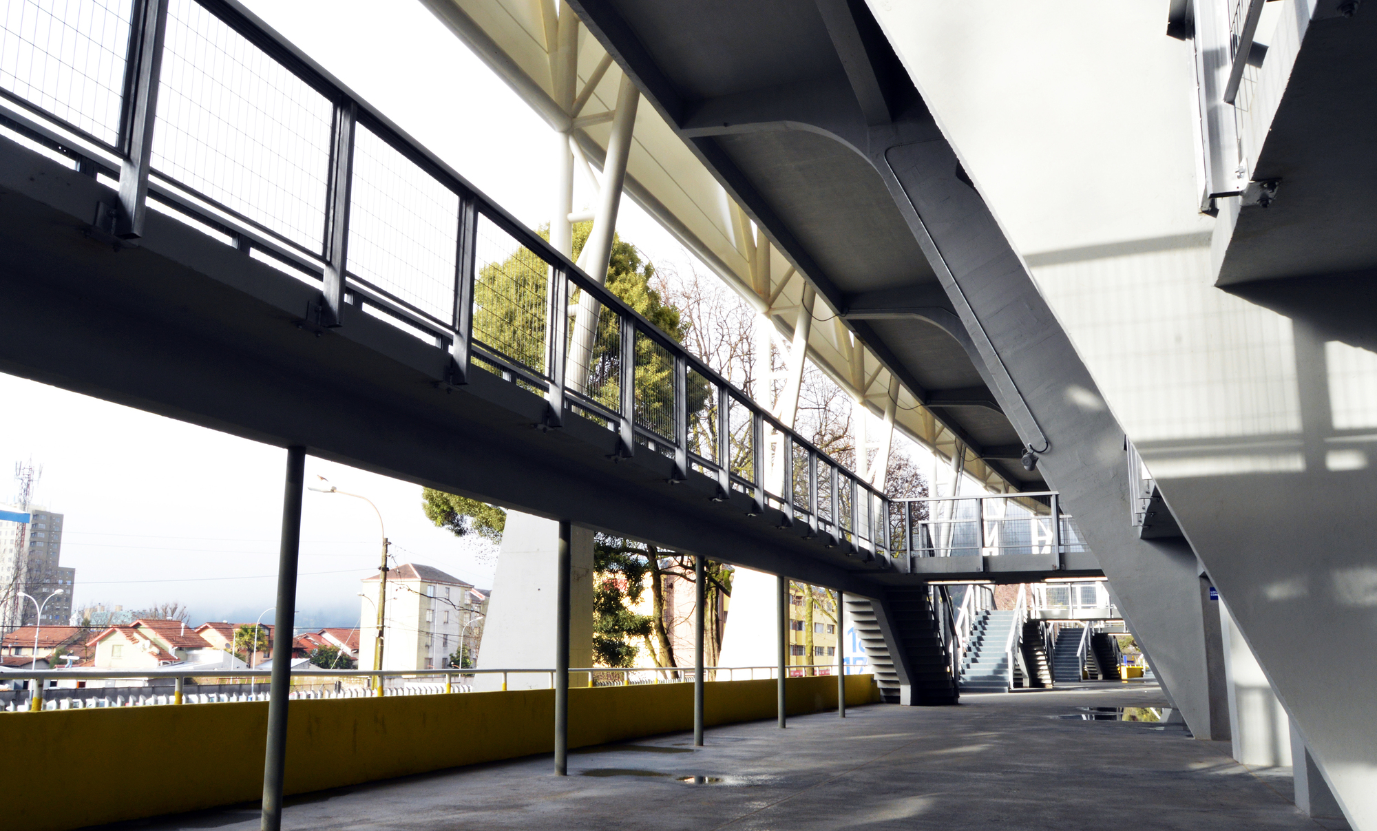 Underside of an elevated concourse walkway with mesh railings, slim steel columns and stairs, lit by winter daylight