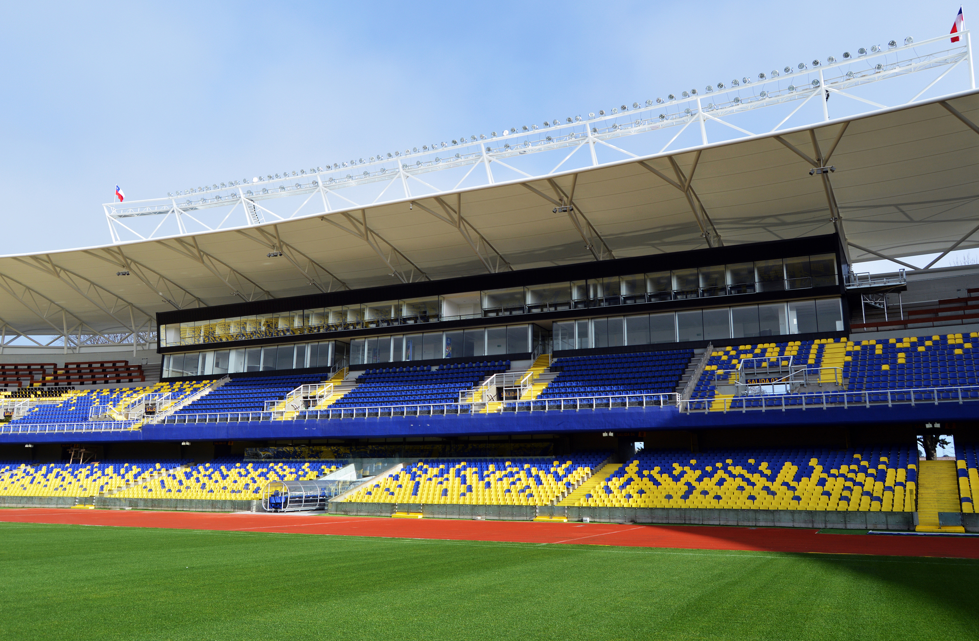 Grandstand with blue and yellow seating, glazed press boxes and a tensile canopy over the pitch on a clear day