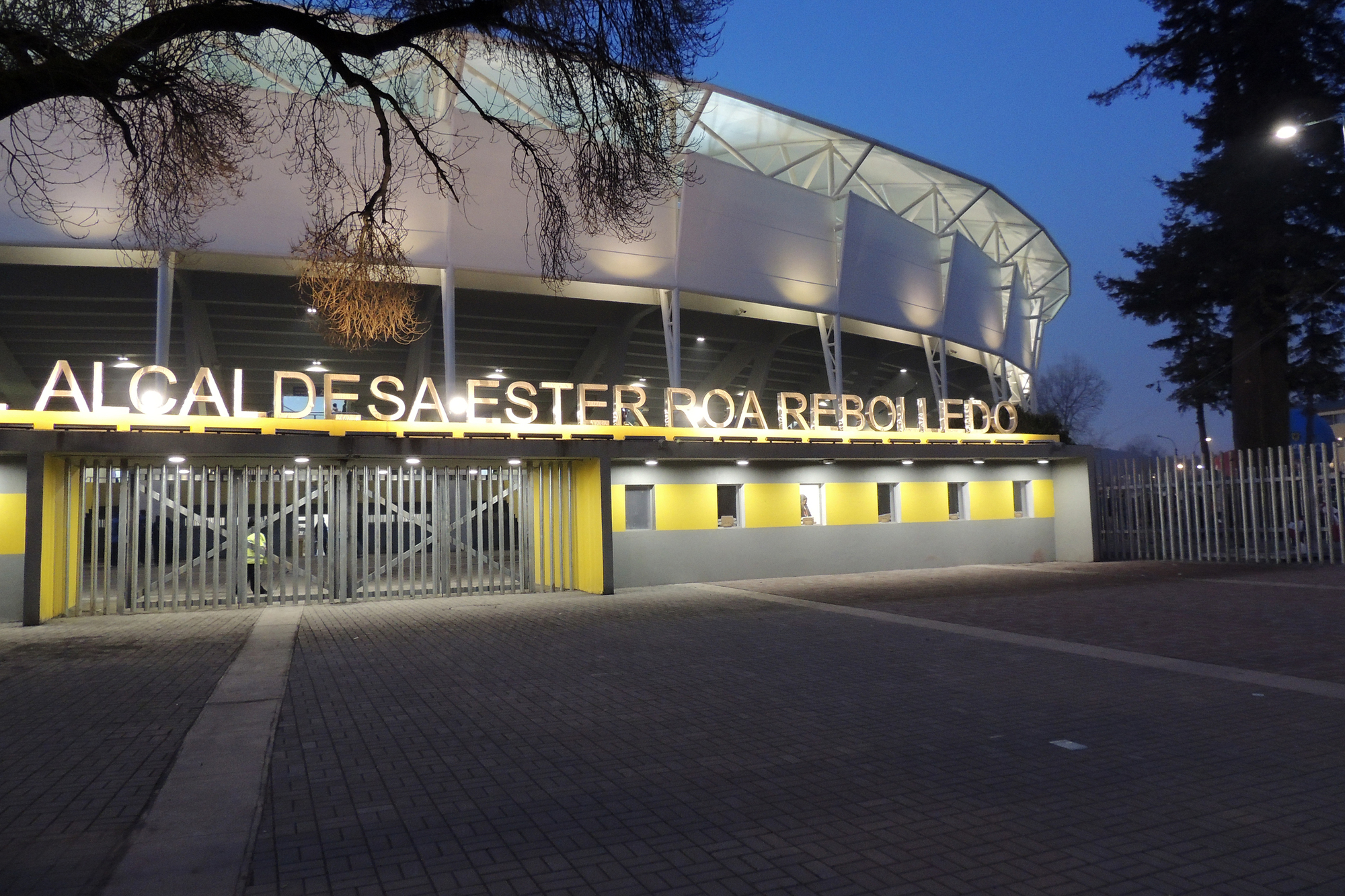 Stadium entrance at dusk with illuminated lettering above yellow-trimmed gates and a bare tree branch overhead