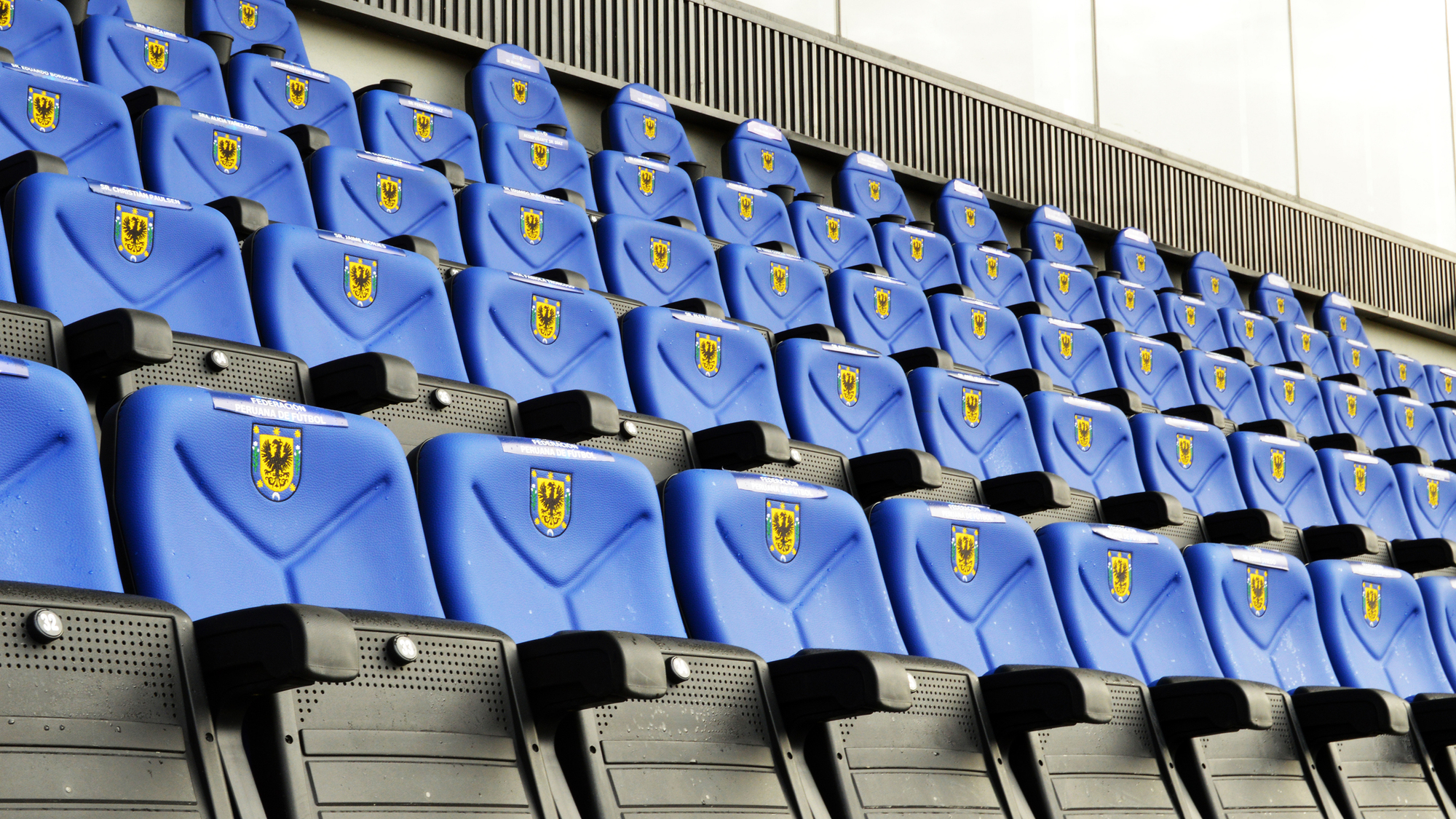 Rows of blue molded seats with yellow crest badges and black armrests beneath a perforated metal band in sunlight