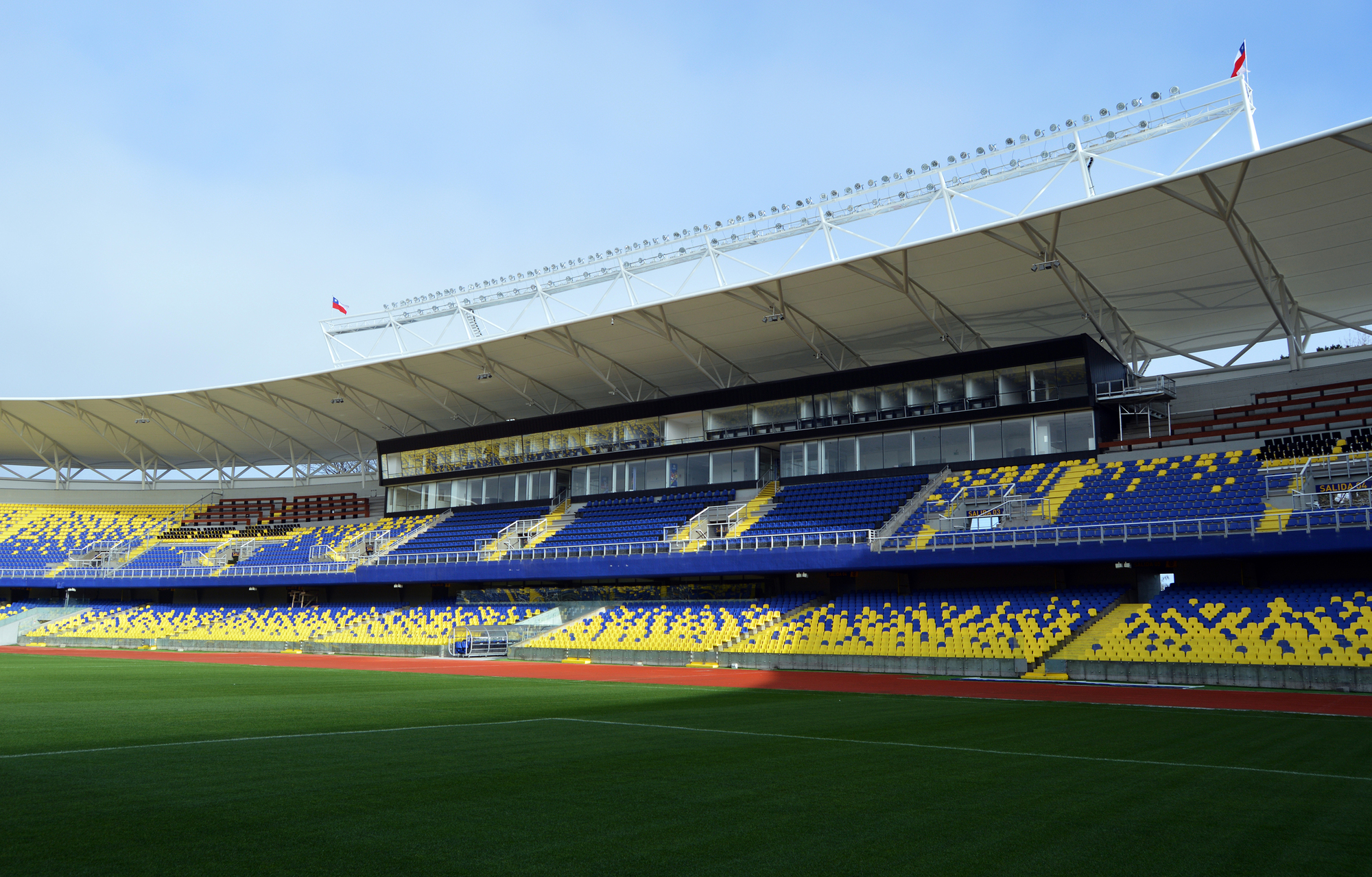 Wide view of the grandstand with blue and yellow seat patterns, glazed boxes and a white canopy above the red running track