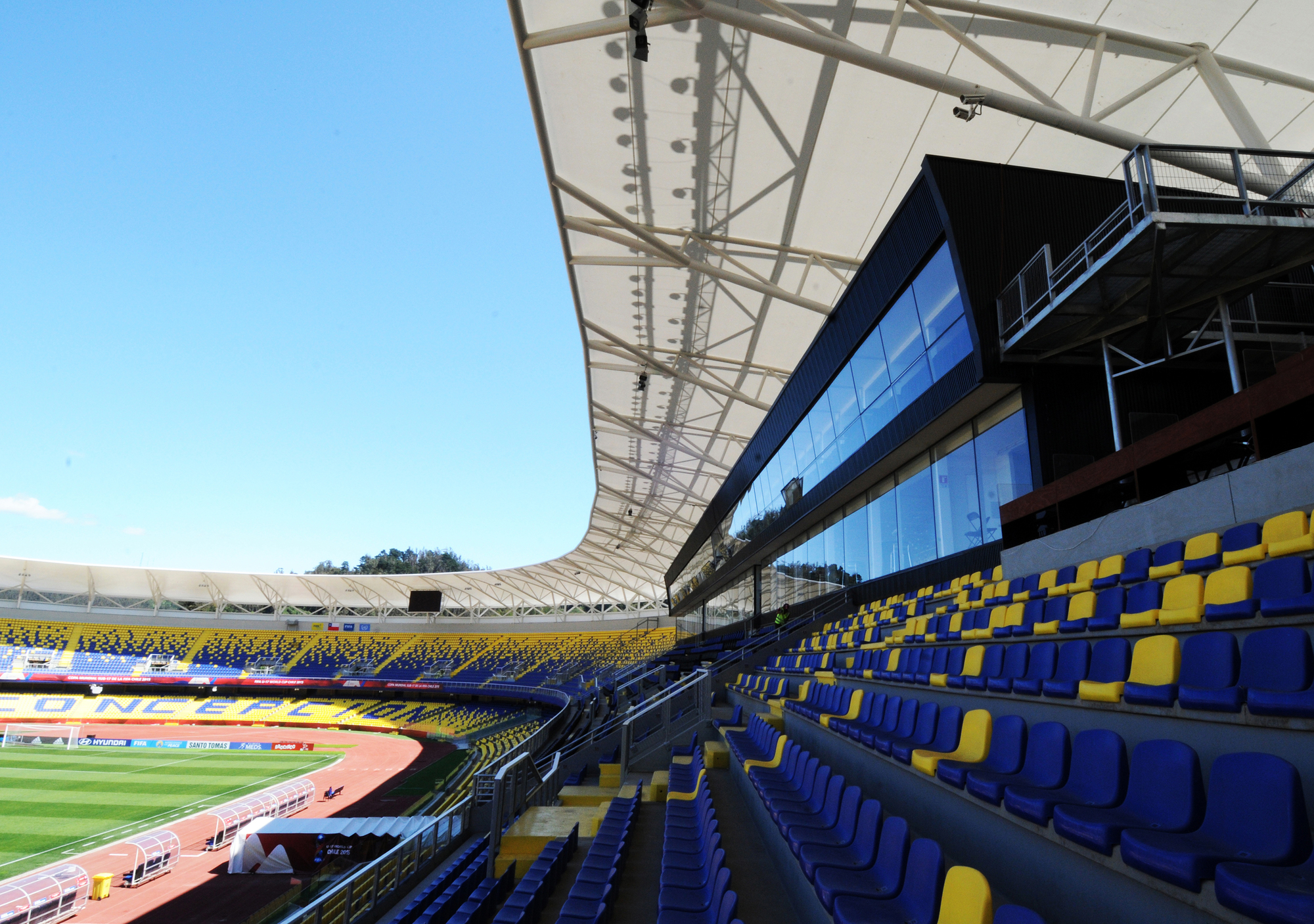 Tensile membrane roof canopy over blue and yellow stadium seating, with a glazed press box beneath a clear blue sky