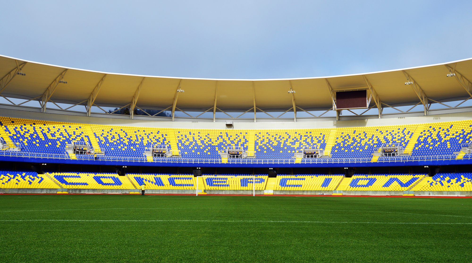 Pitch-level view of the curved stadium roof edge above blue and yellow seat tiers under a pale sky