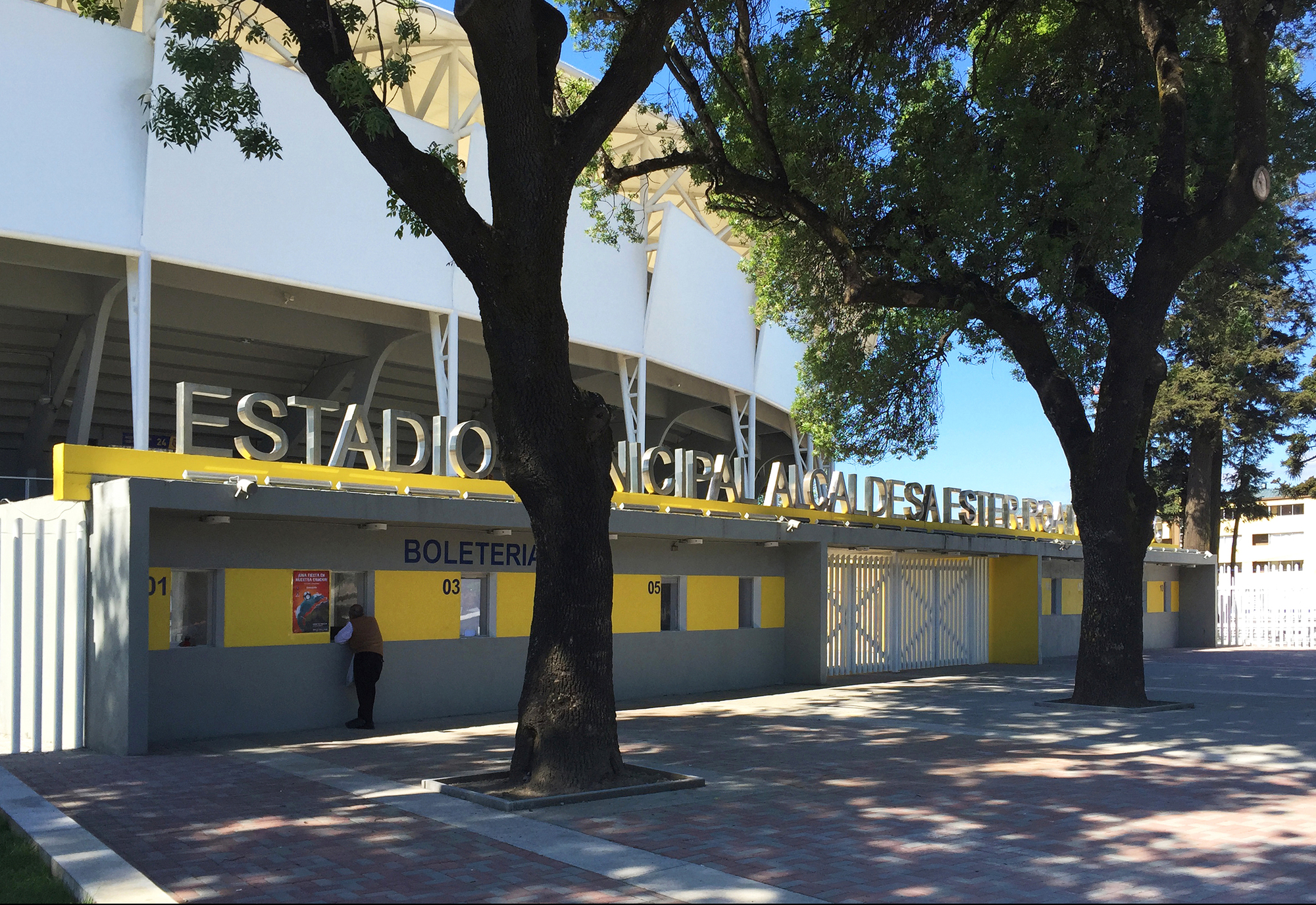 Ticket office frontage with yellow band and lettered signage, shaded by two large trees, a person standing at a window