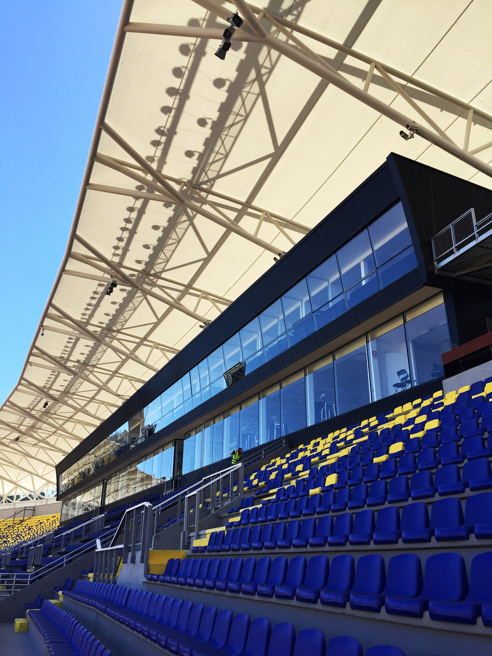 Low angle of the white membrane roof and steel trusses above a dark-clad glazed box and blue seating rows
