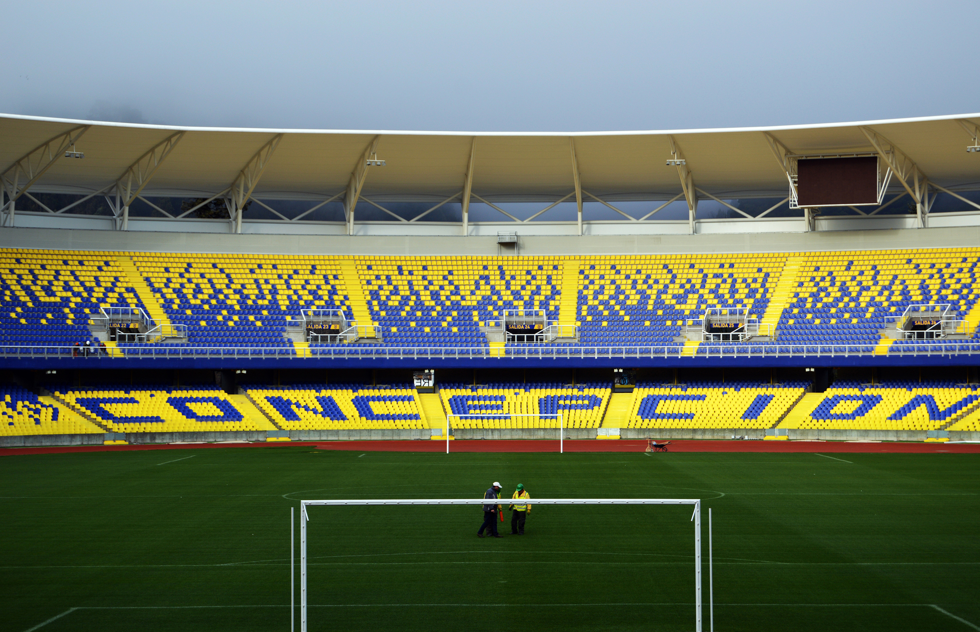 Two workers stand near a goalpost on the grass, facing the tiered blue and yellow stands under an overcast sky