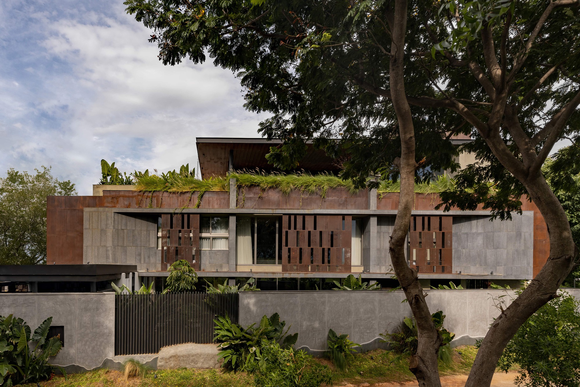 Weathered steel and grey stone facade with perforated panels and a planted roof edge, seen through tree trunks under a cloudy sky