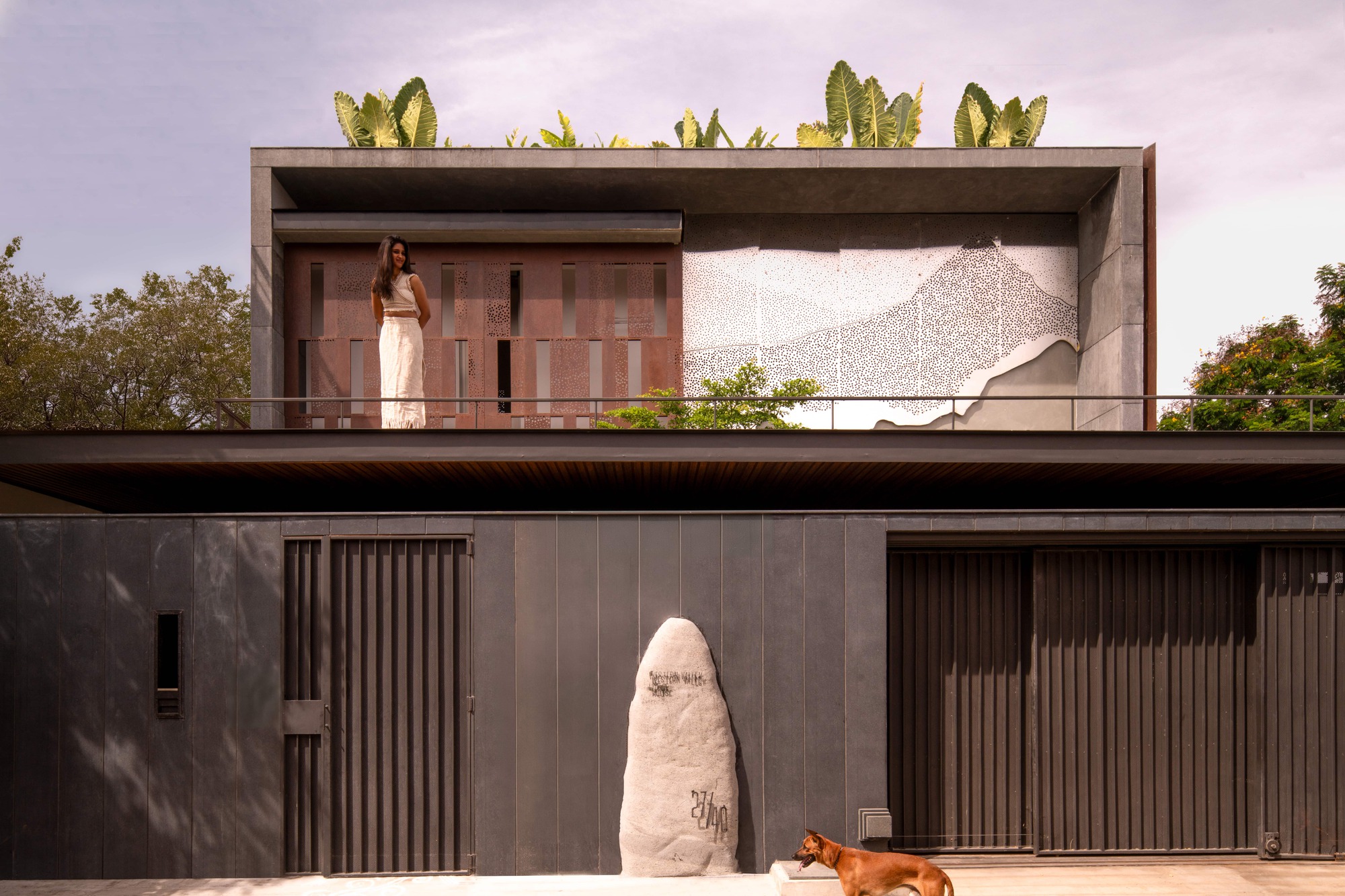 Street facade with dark vertical metal gates, a standing stone and a perforated mountain-patterned panel above, with a dog in the foreground