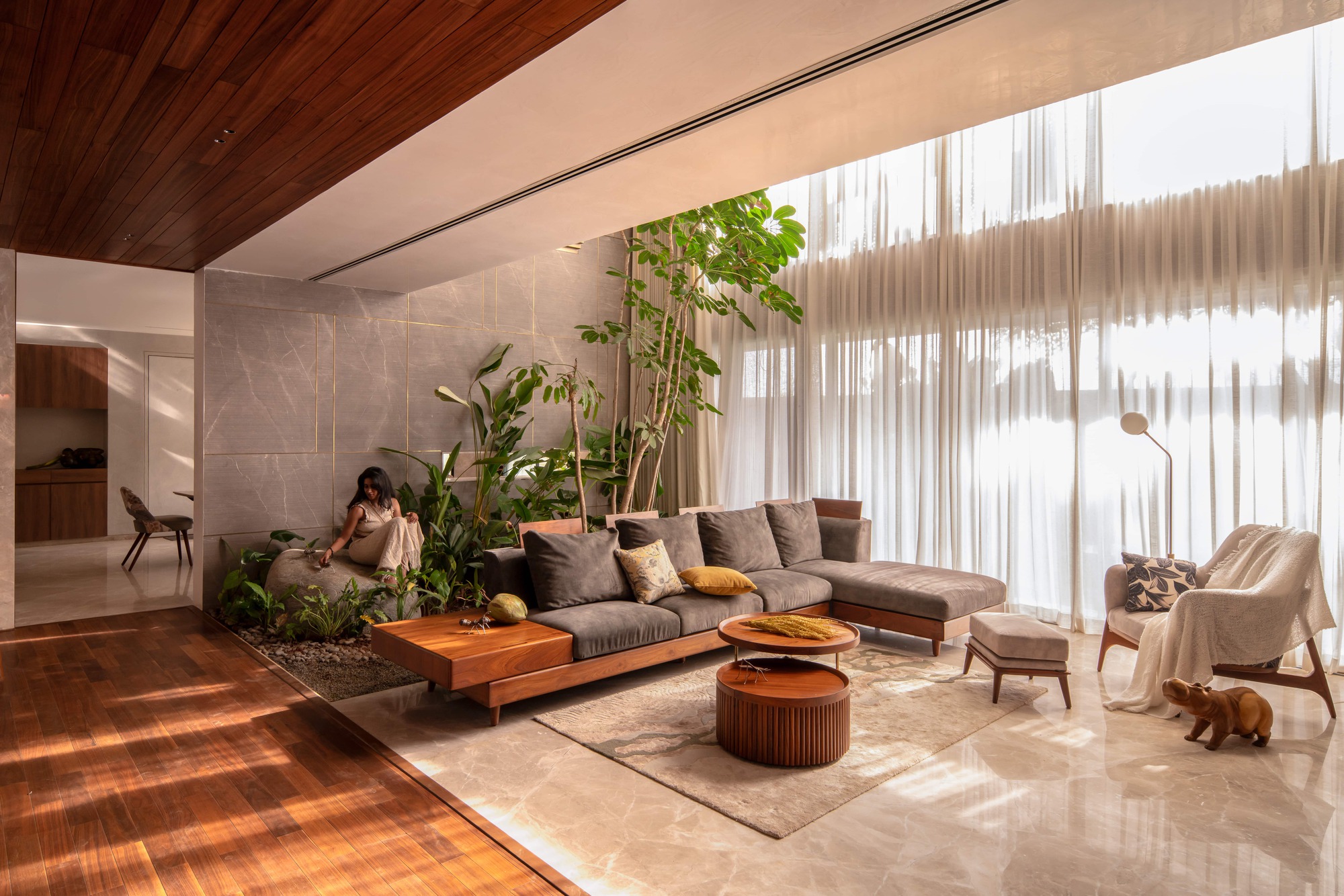Living room with a low timber-framed sofa, indoor plants and floor-to-ceiling sheer curtains, sunlight falling across the wood floor