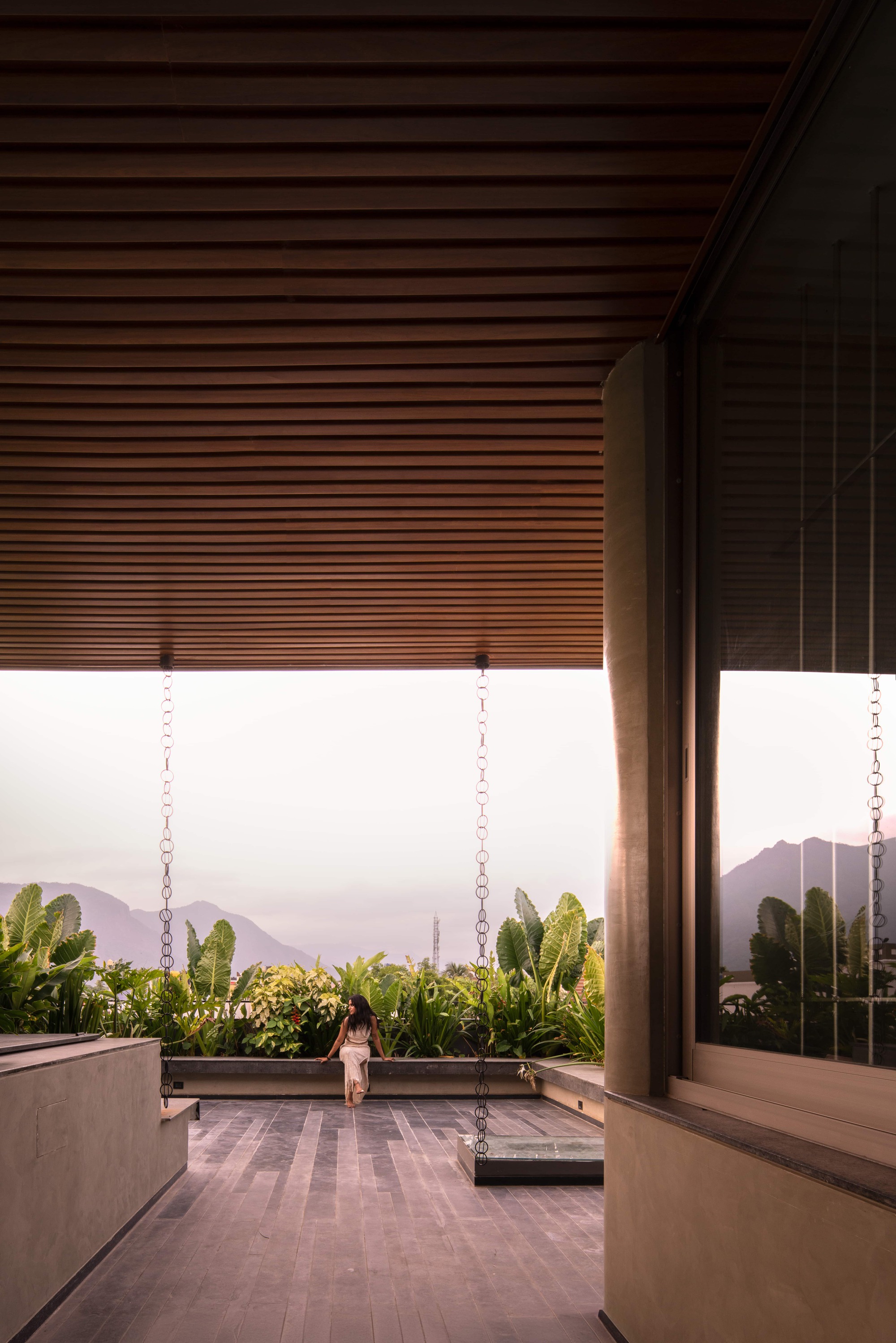 Covered terrace with slatted timber ceiling and hanging rain chains, a seated figure facing planting and misty hills at dusk
