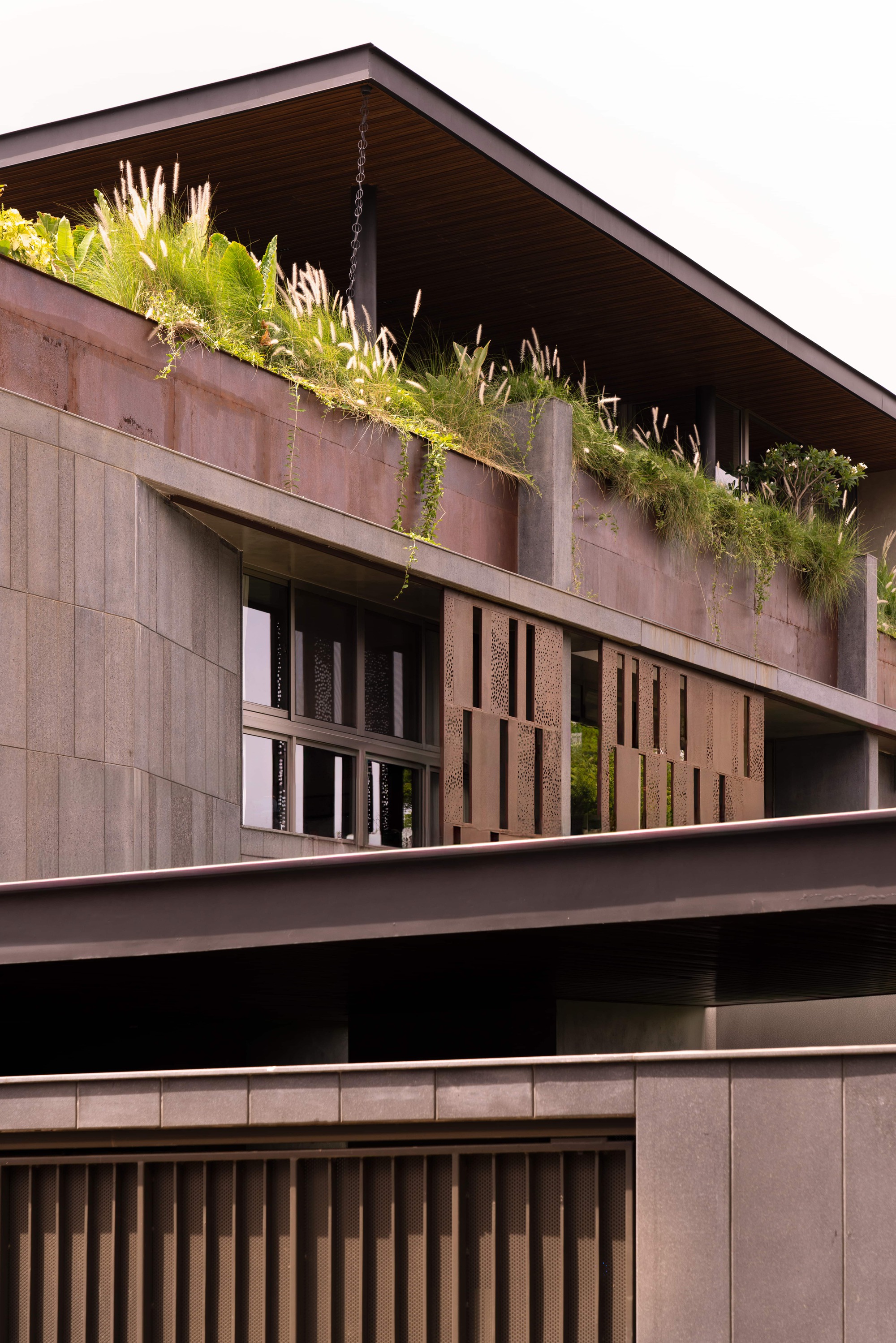 Upper floors with a planted rooftop edge of grasses above weathered steel panels, perforated screens and stone cladding in soft daylight