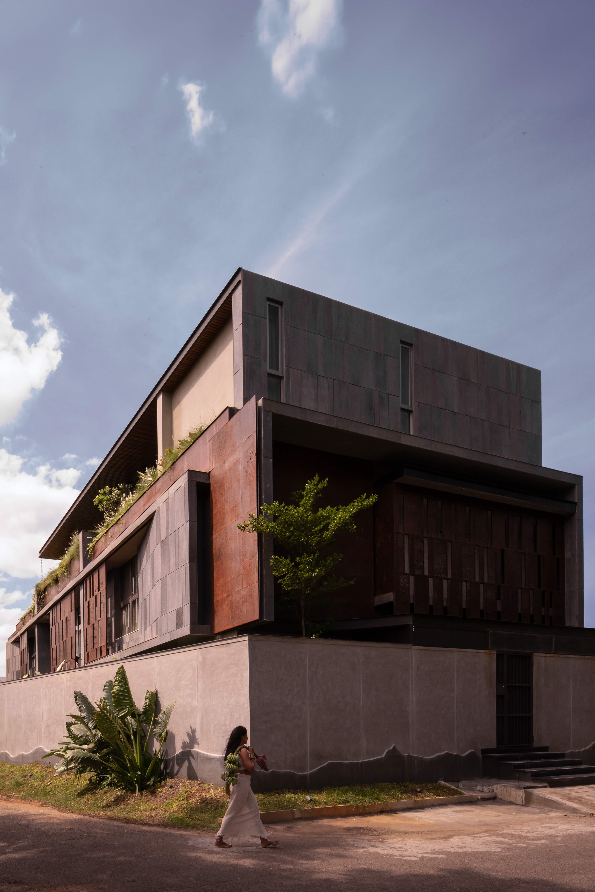 Corner of the stepped stone and rust-toned steel volume above a boundary wall, with a woman walking past under scattered clouds