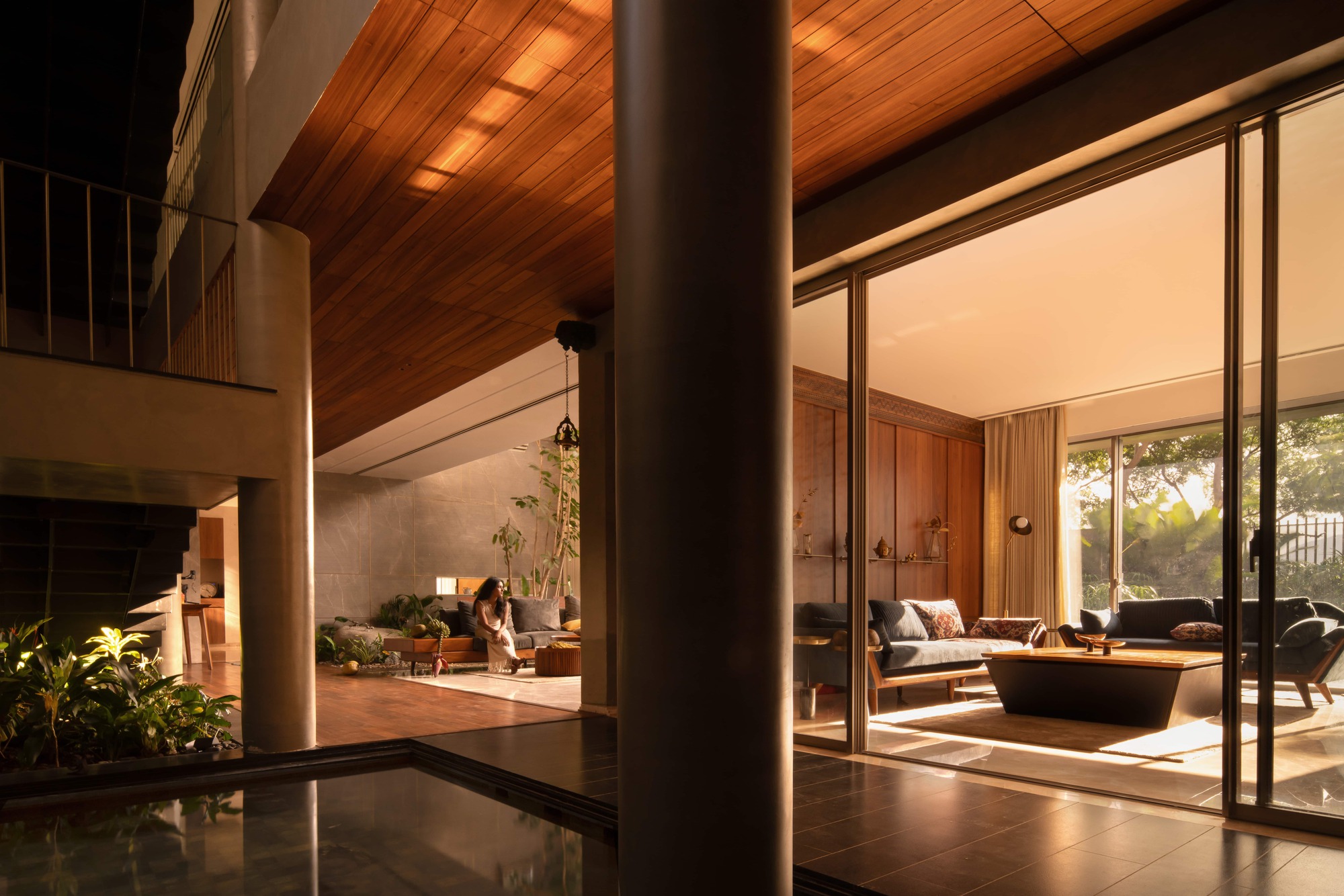Timber-ceilinged living space seen past a column and reflecting pool, with low sunlight falling across sofas and a seated woman