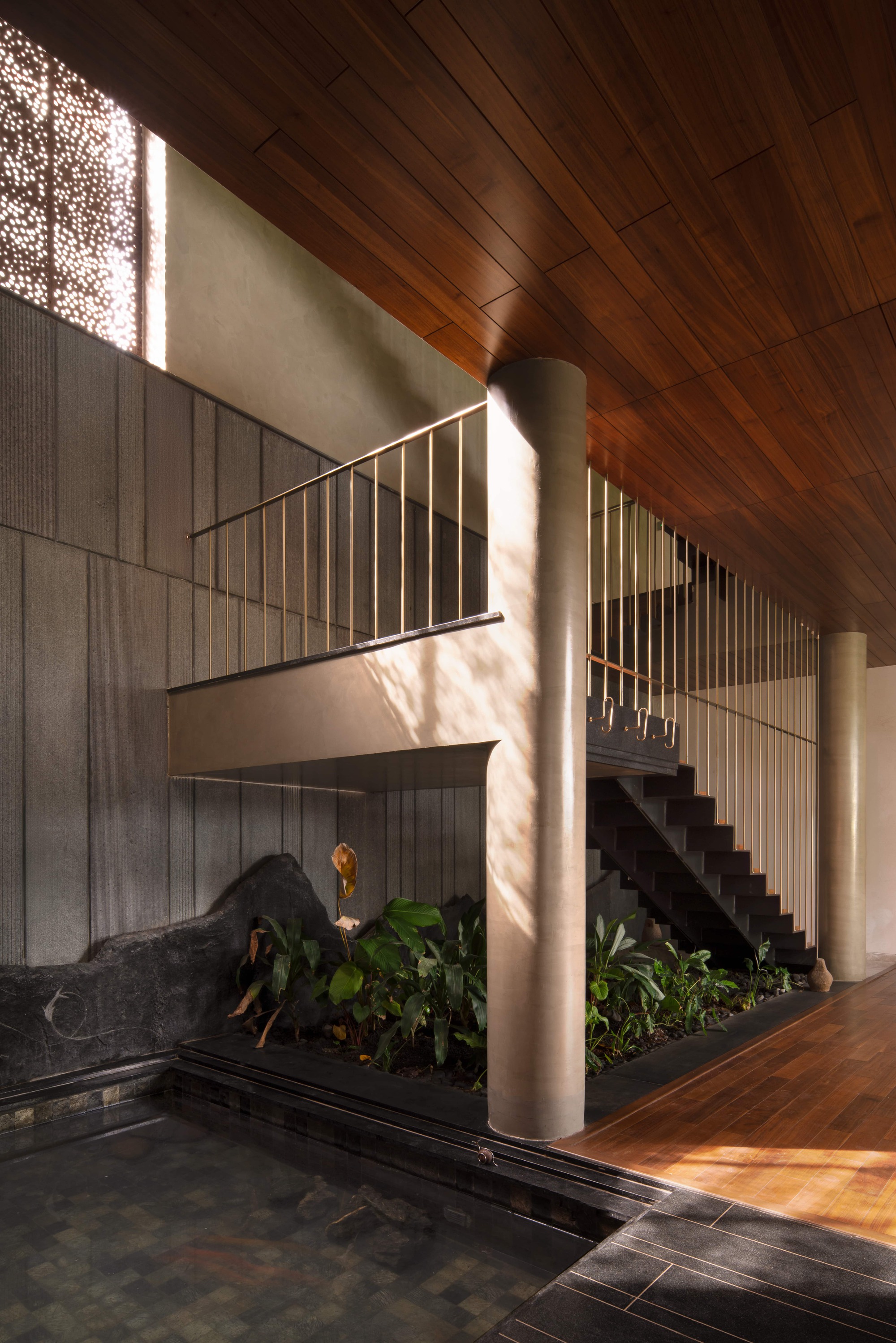 Staircase with slim brass balusters beside a round column, a planted bed and a dark water pool under a timber ceiling