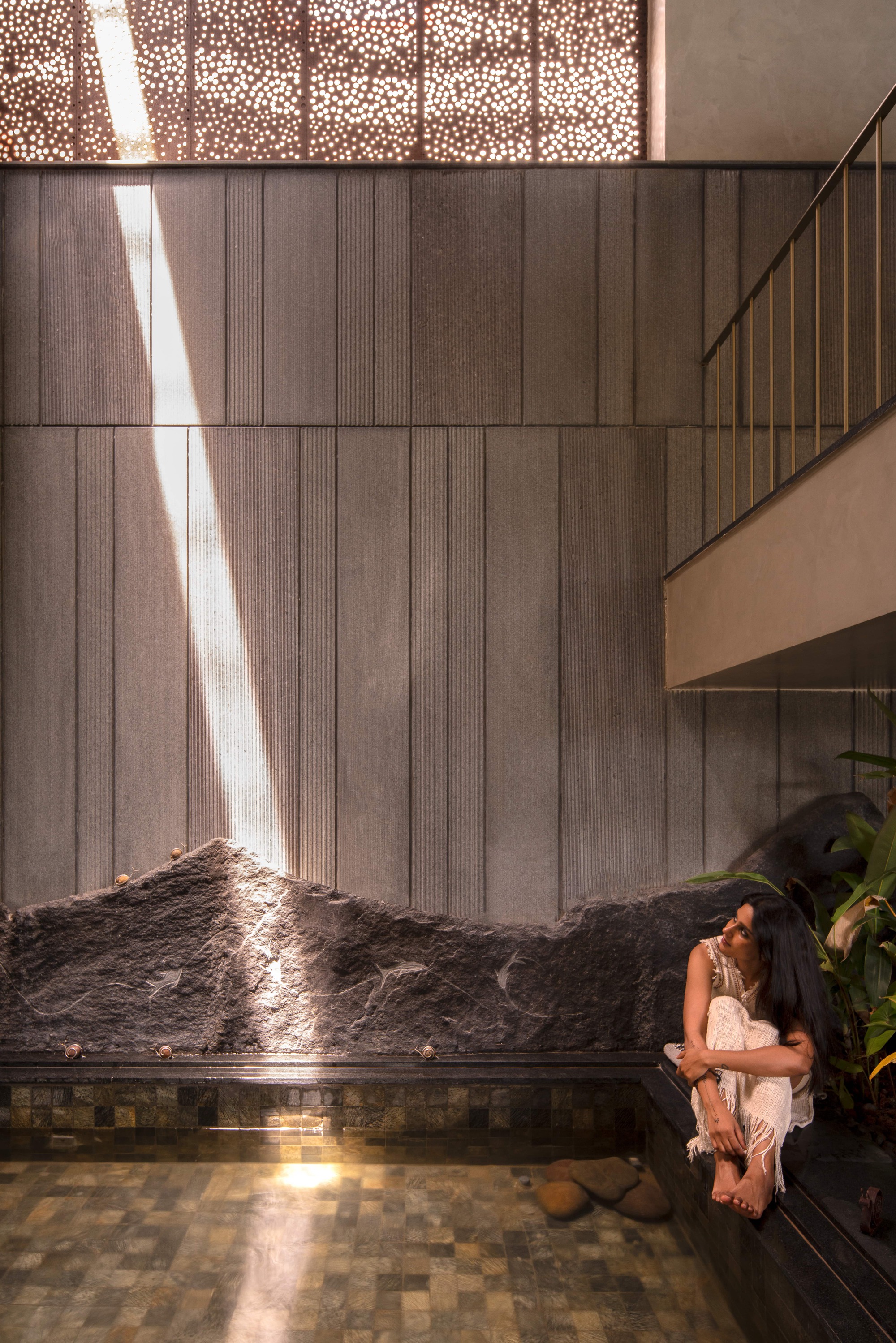 Shaft of sunlight from a perforated screen crossing a ribbed stone wall above a rough rock edge, with a woman seated at the pool