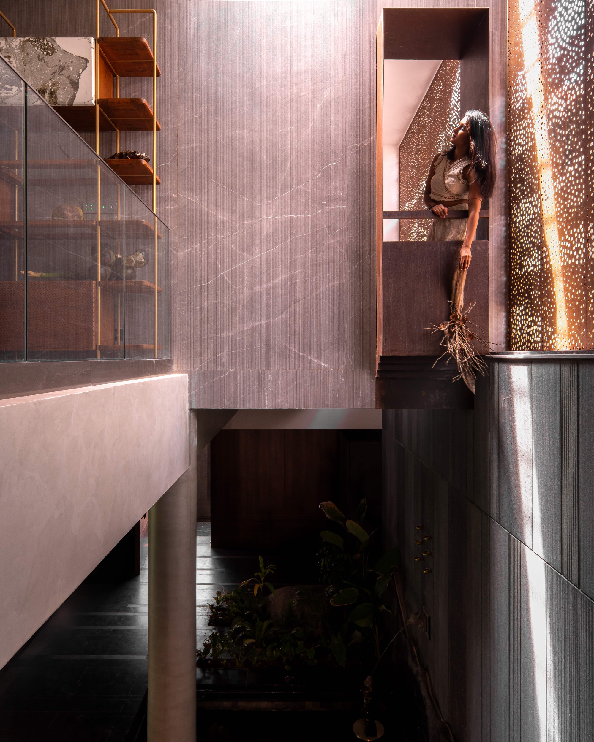 Double-height void with pink marble walls, a perforated screen and a woman leaning from an upper balcony above a planted court